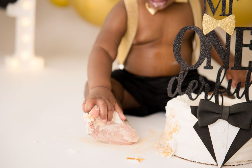 A small hand reaches for a cake during a first birthday celebration with black and gold decorations.