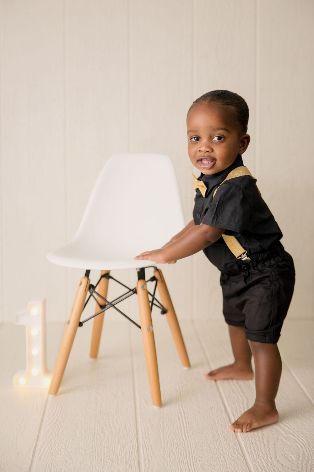 Baby in formal outfit standing beside modern white chair during photo session.