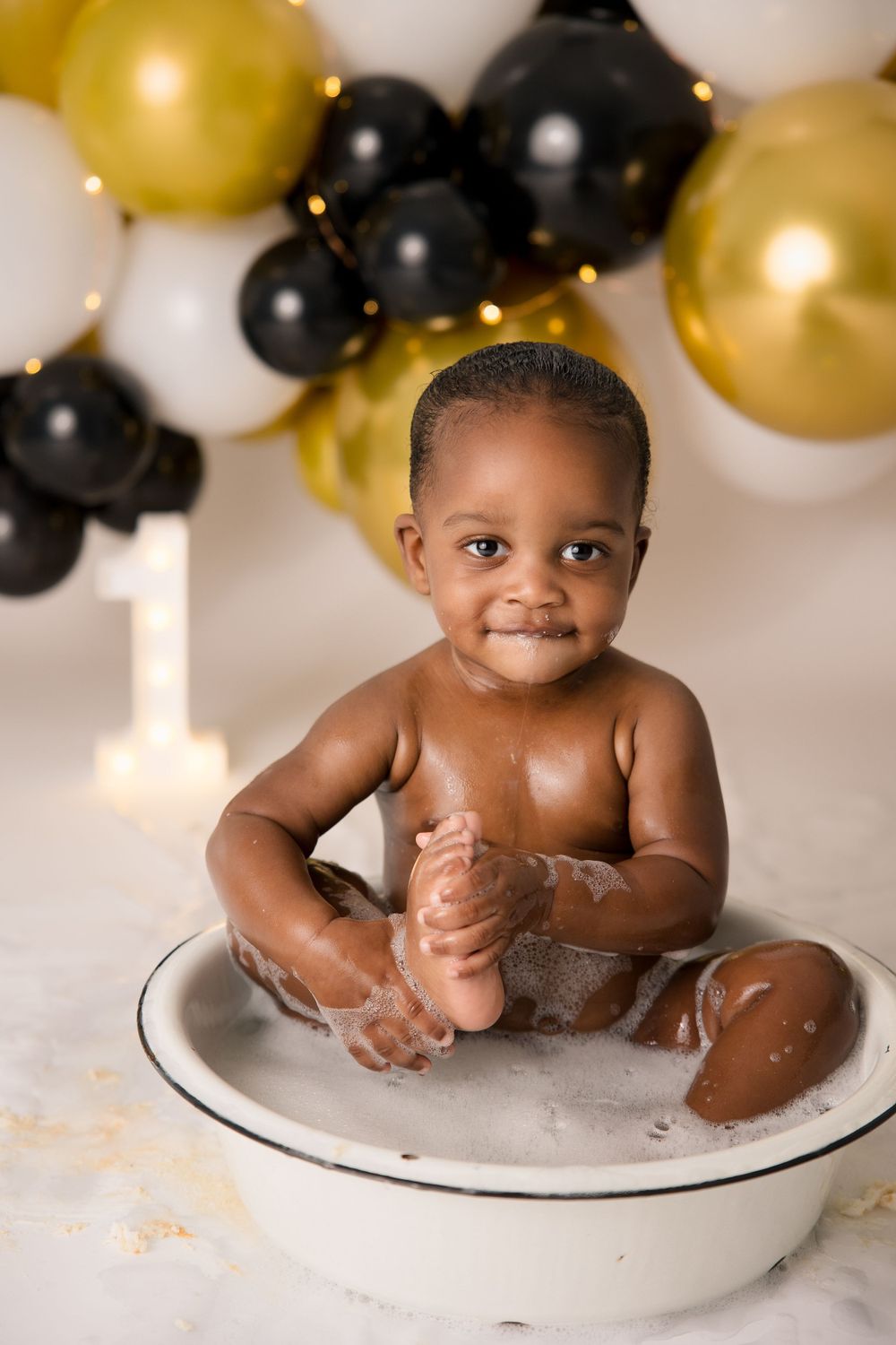 Smiling baby plays in a white basin with soap during a first birthday photoshoot.