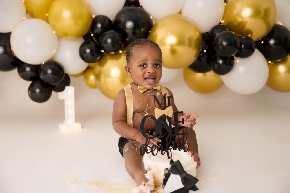 Toddler sits among black and gold party balloons during a festive photoshoot.