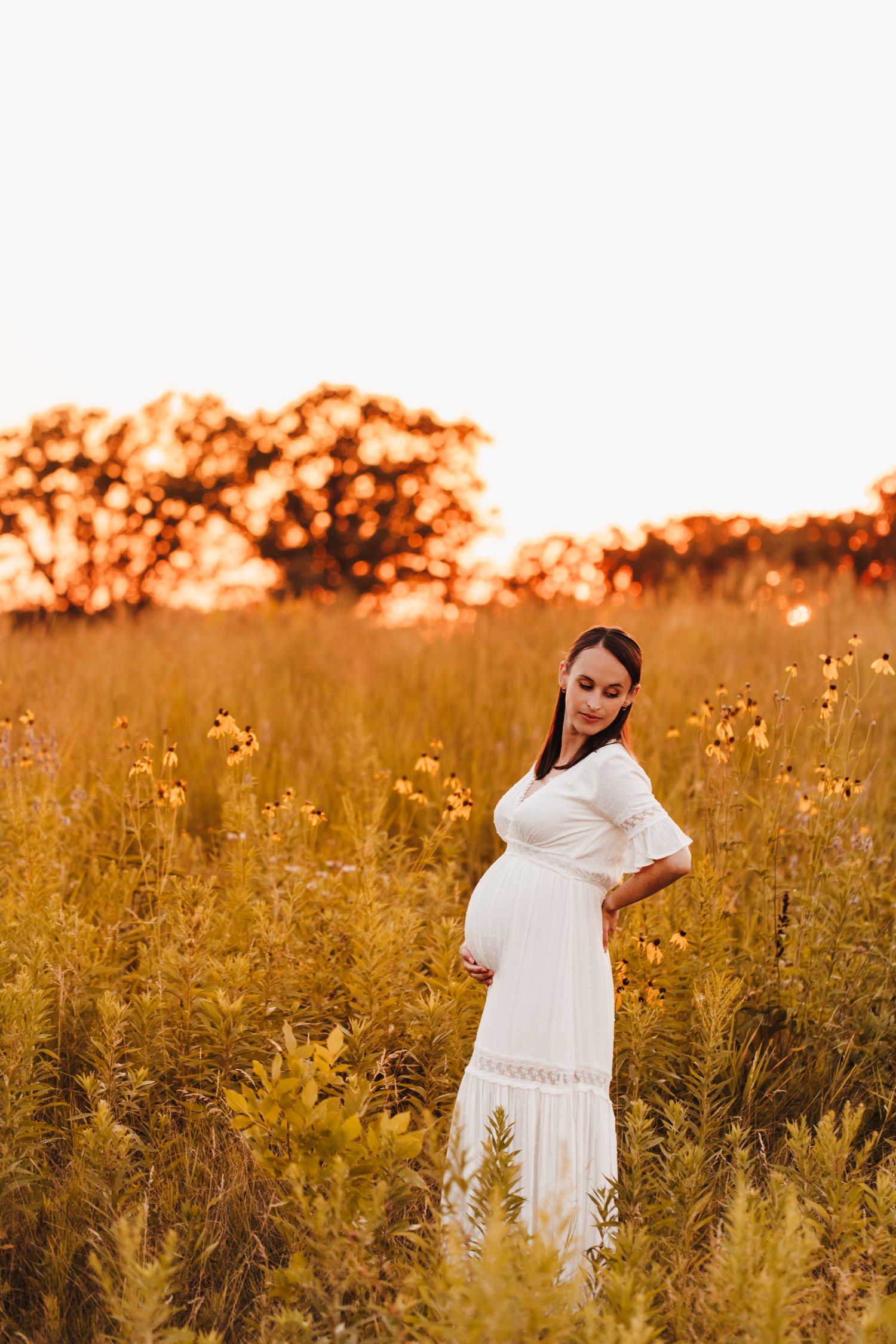 Pregnant woman in a white textured dress stands in a field of wildflowers during golden hour
