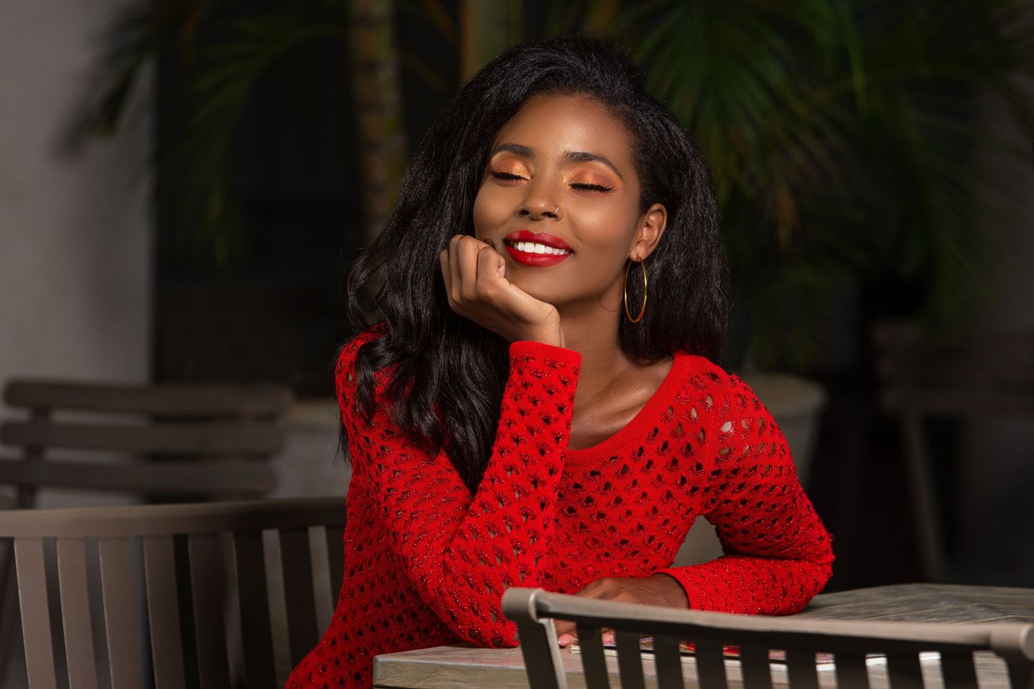 Woman in red dress smiling while sitting at outdoor table with dark hair and bright lipstick.