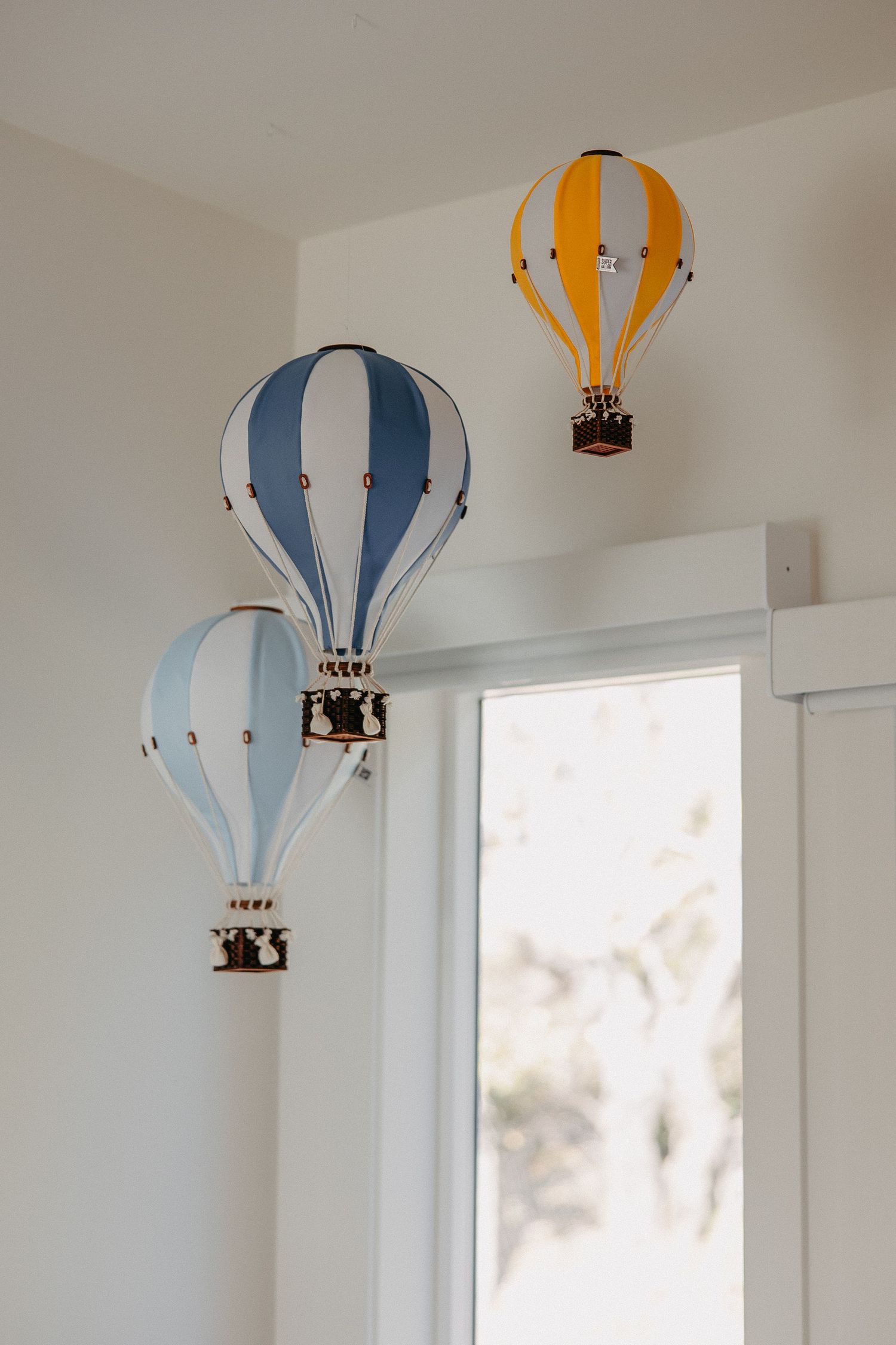 Three decorative hot air balloon ornaments in blue, white and yellow mounted on a white wall near a window.