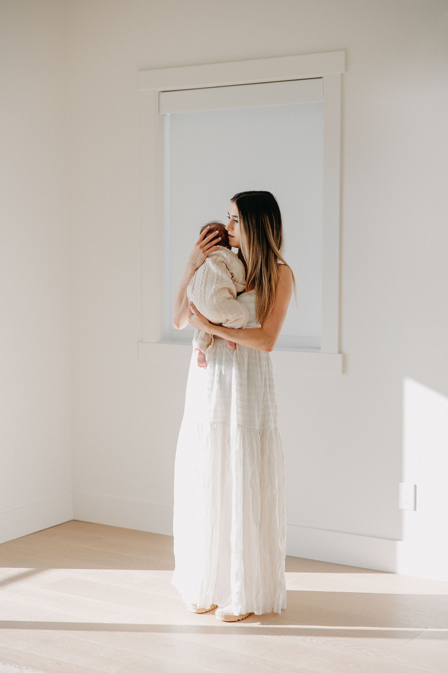 Woman in flowing white dress holds baby while standing near window in bright minimalist room.
