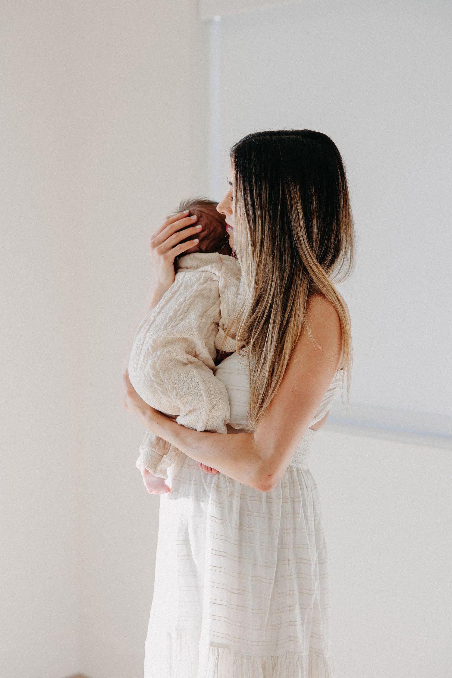 Woman in white dress holding baby in cream-colored outfit against white wall in minimalist setting.