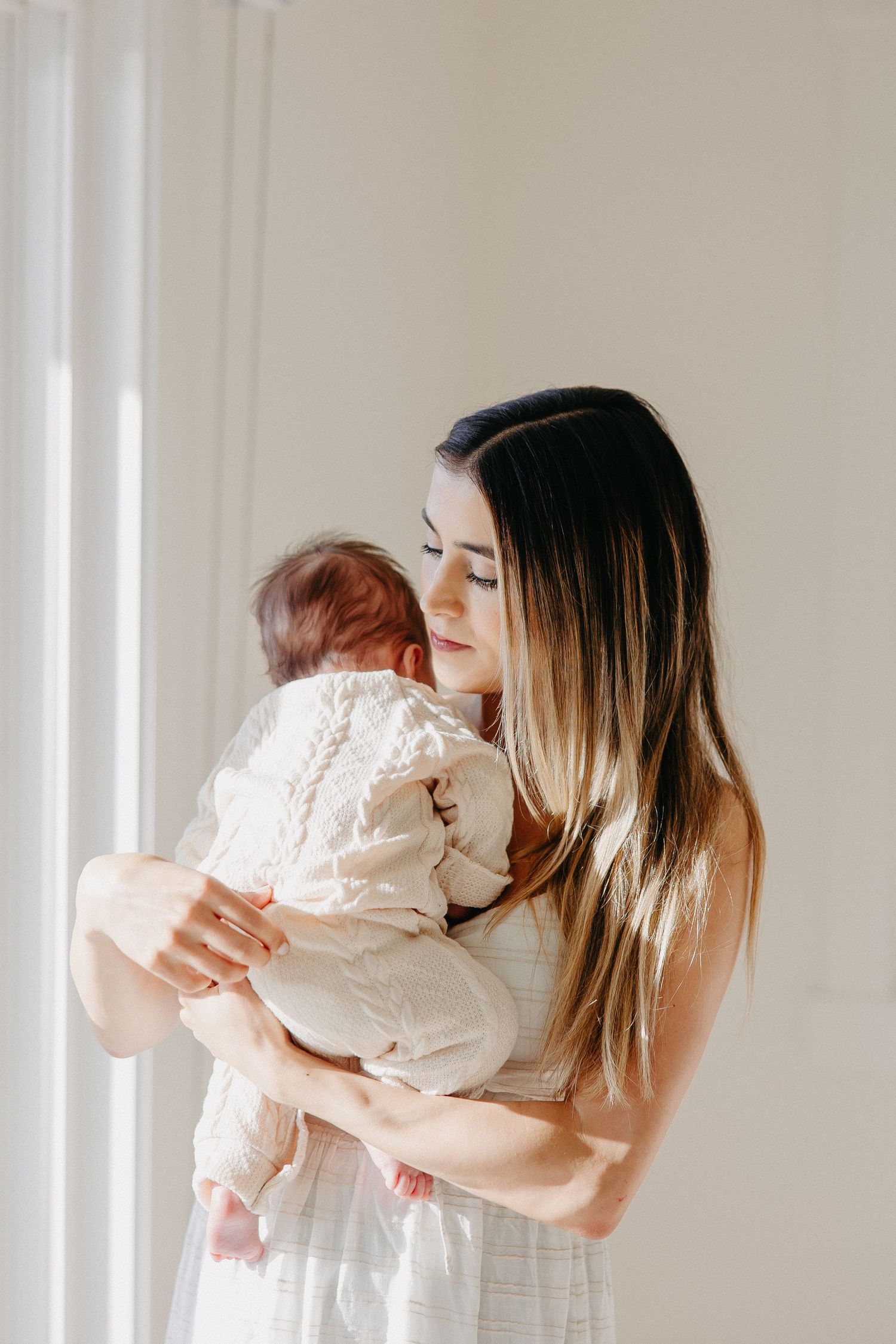 Intimate moment between parent and baby in white clothing by sunlit window.