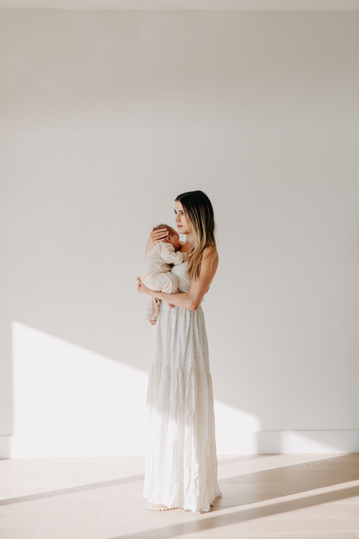 Minimalist portrait of a person in white maxi dress holding a baby in a sunlit room.