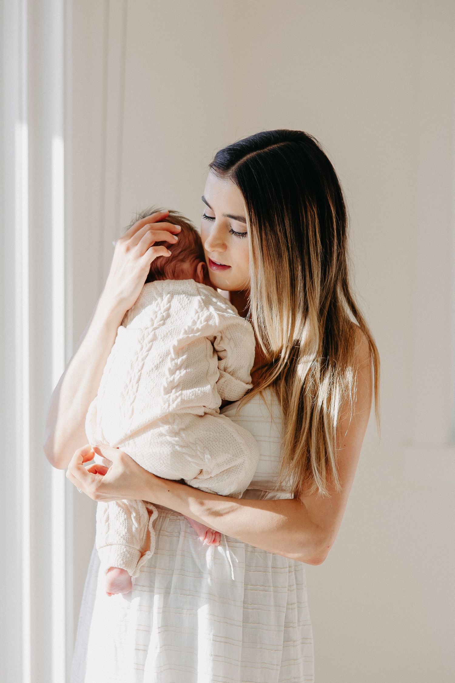 Tender embrace between mother and baby in matching white outfits by bright window.