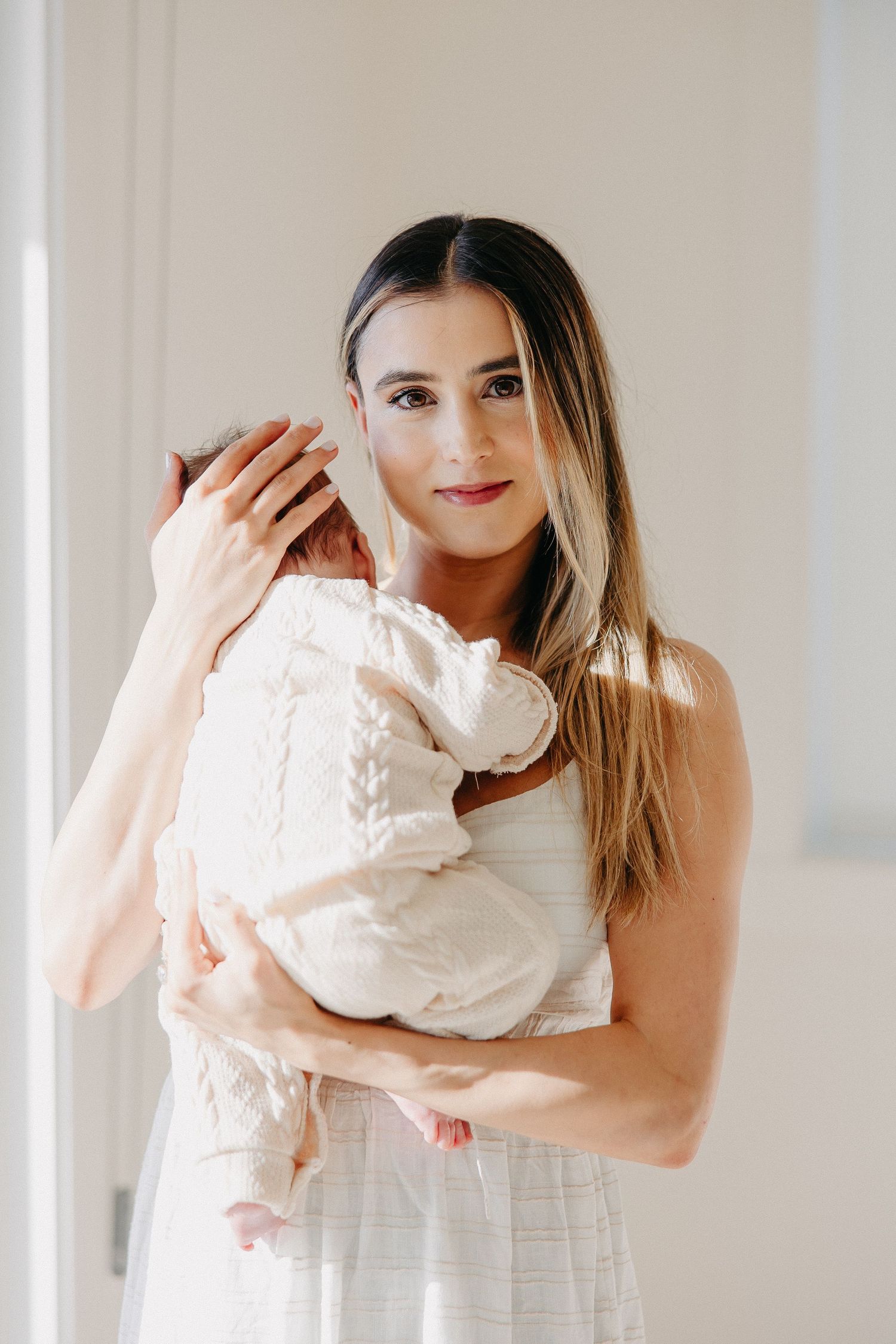 Portrait of woman in white dress holding baby against bright interior wall.