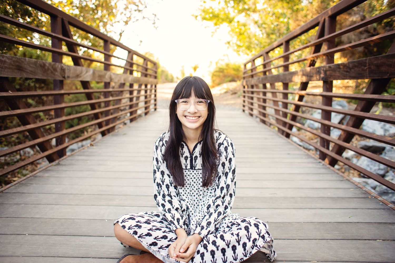 Person sitting cross-legged on a wooden bridge in a patterned dress during golden hour.