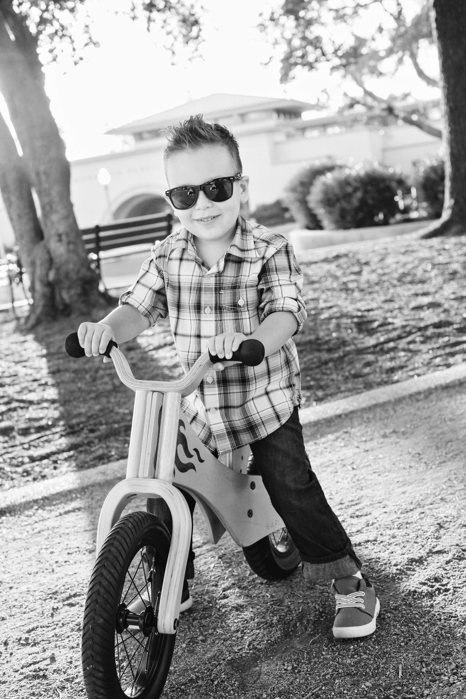Black and white photo of young person in plaid shirt and sunglasses on balance bike.