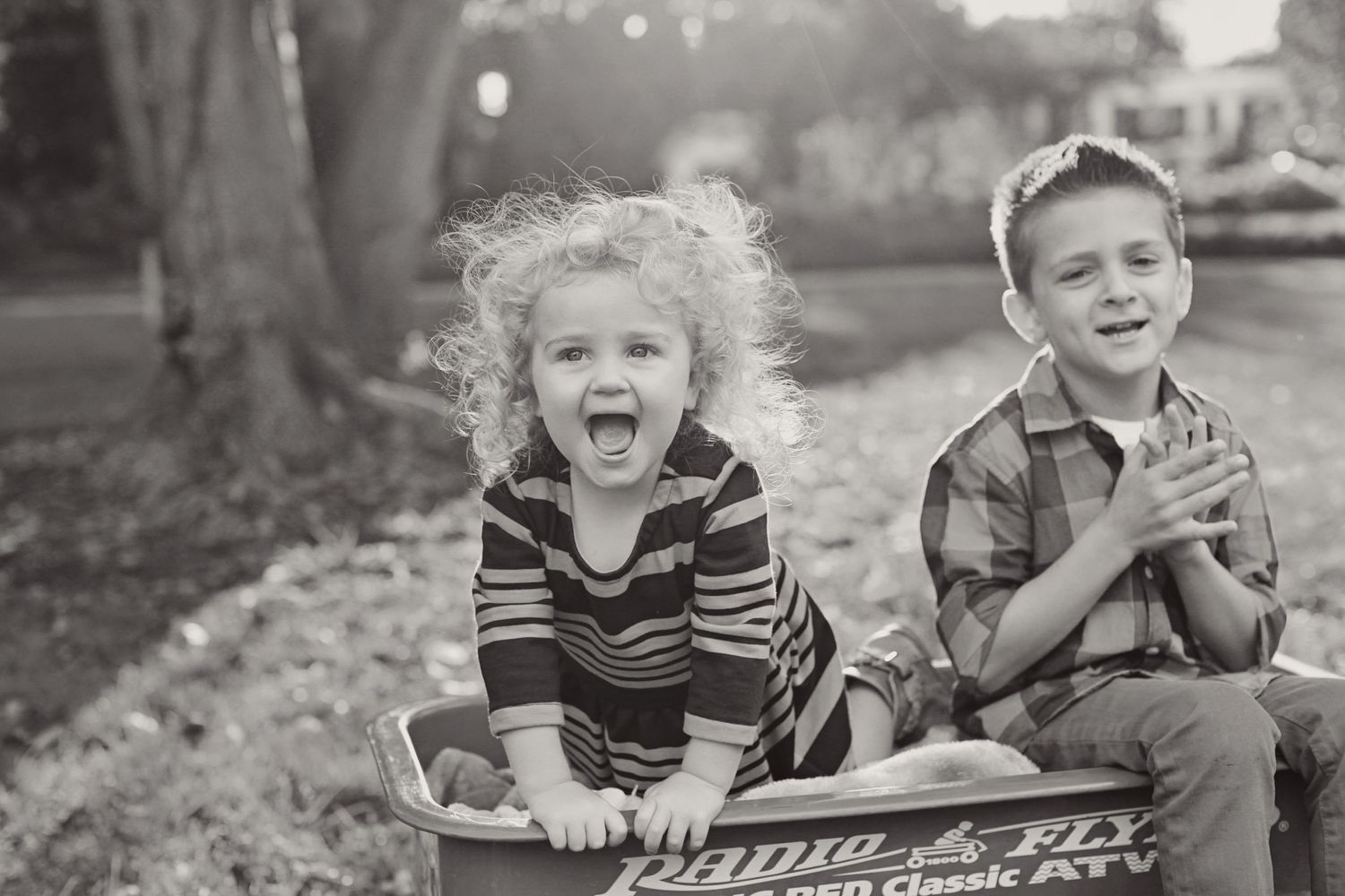 Black and white photo of two children sitting on vintage wagon outdoors.
