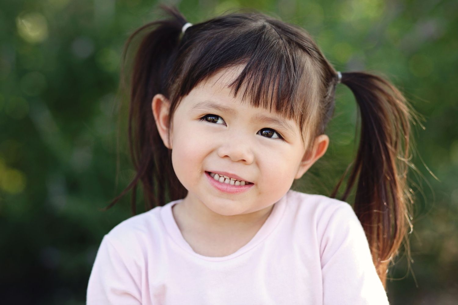 Child in pink shirt with pigtails smiles against blurred green background.