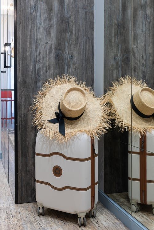 Two straw sun hats resting on a white suitcase against a wooden wall with a mirror reflection.
