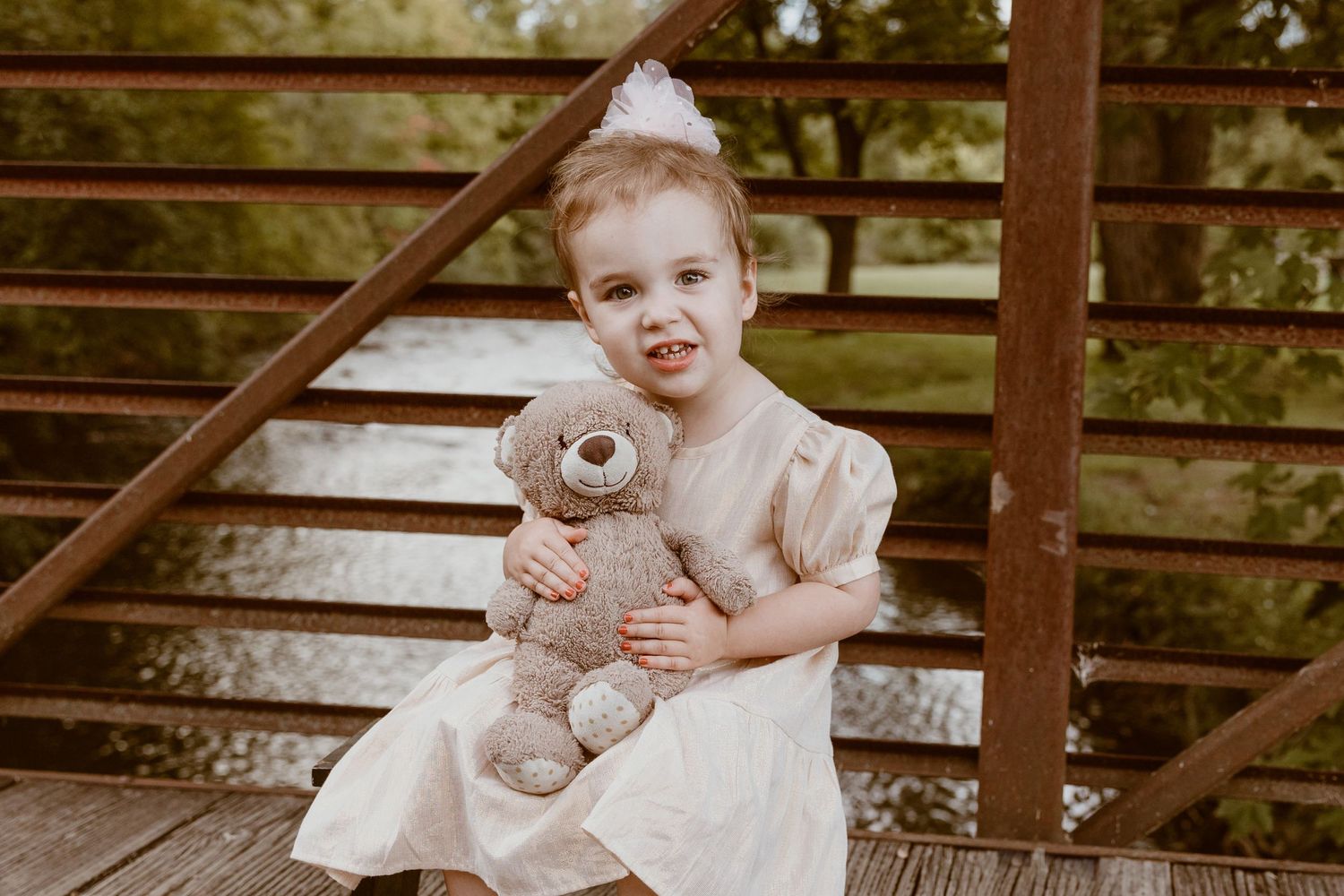 A small child in a light dress hugs a teddy bear while sitting on wooden steps outdoors.