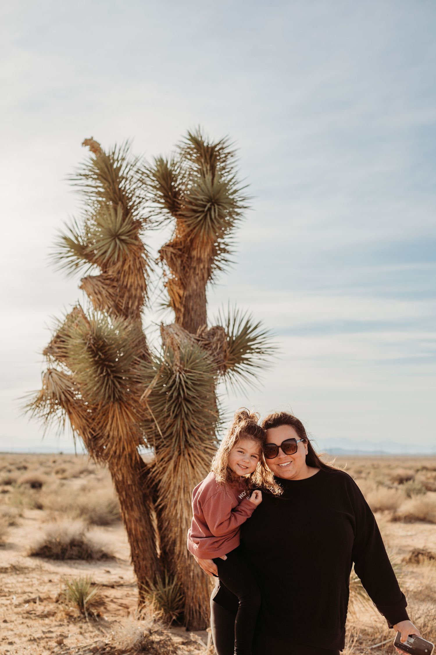 A couple poses together in front of a tall Joshua Tree in a desert landscape during sunset.
