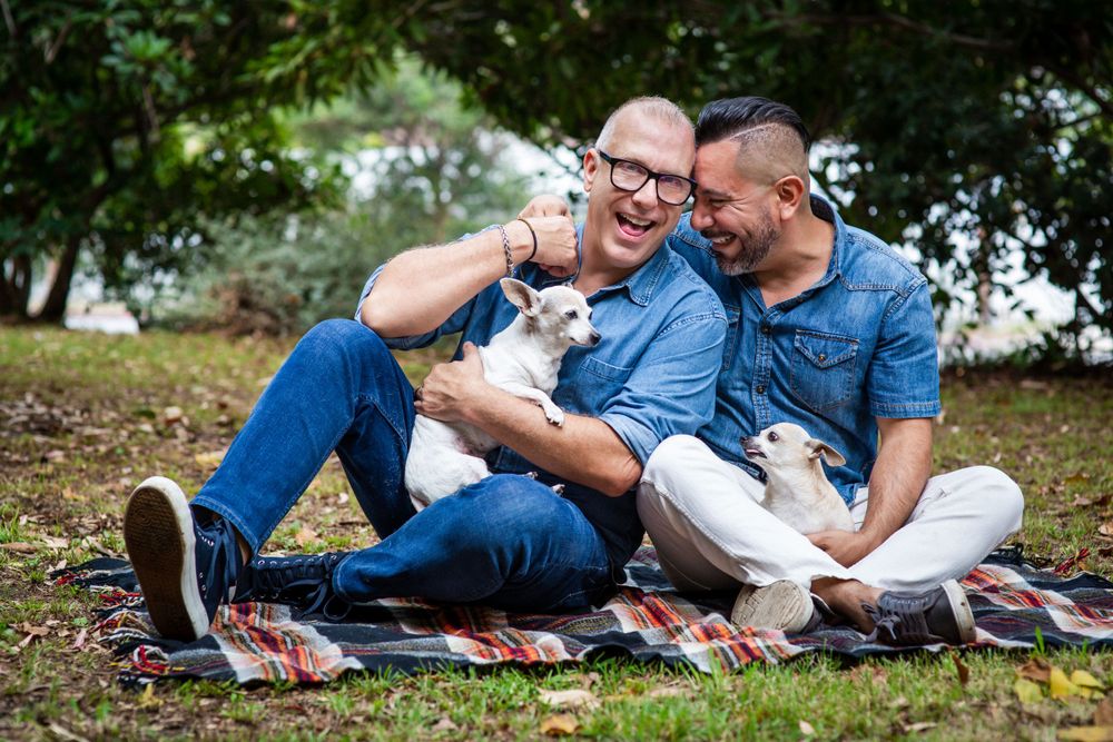 Two people laughing together on a picnic blanket while playing with their small white dog.