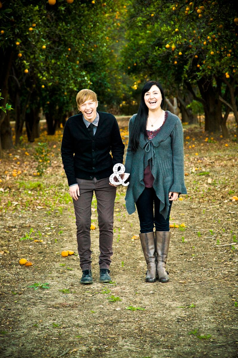 A couple stands in an orange grove with an ampersand symbol between them during their engagement shoot.
