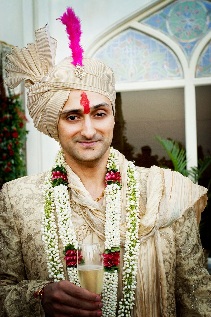 A groom in traditional Indian wedding attire with floral garlands and a decorative turban holds a drink.