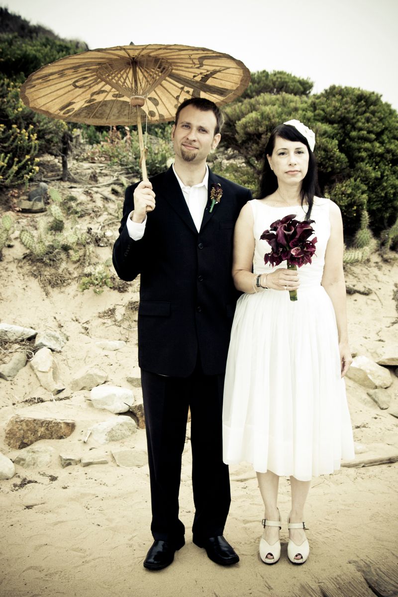 A couple poses on a beach with a paper parasol and burgundy bouquet in vintage-style wedding attire.