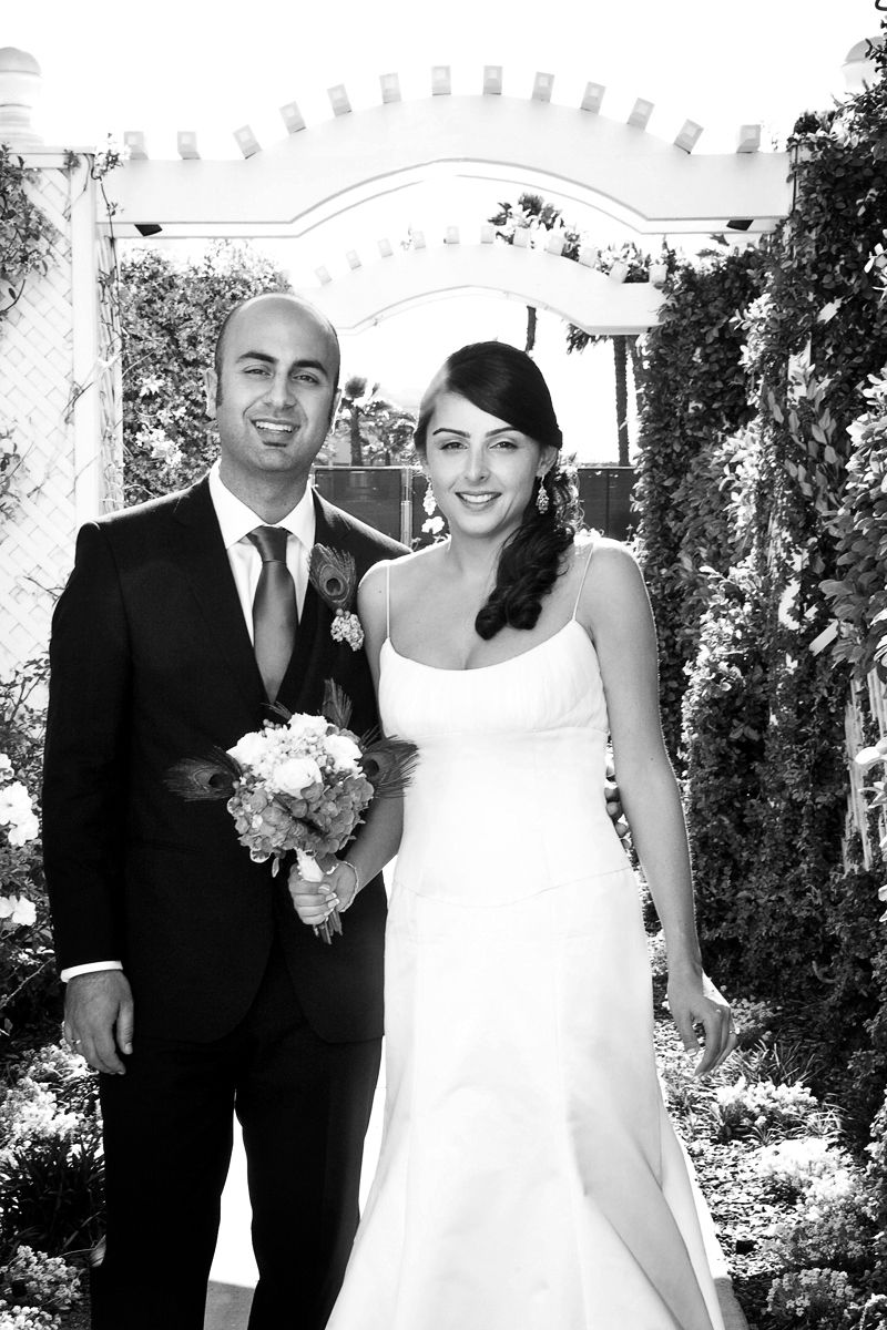 A wedding portrait under a white garden arch with greenery and flowers in black and white.
