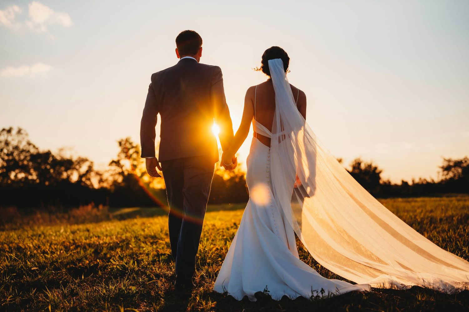 Silhouette of newlyweds walking at sunset with flowing wedding dress veil.