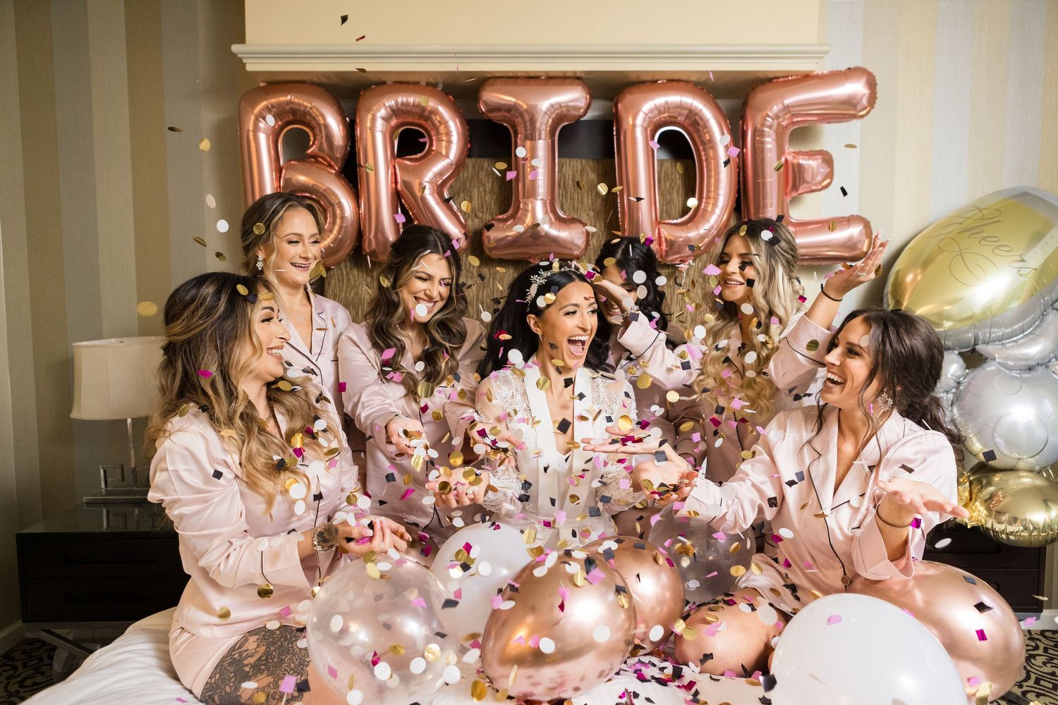 Rose gold BRIDE balloons decorate a room while pink confetti fills the air during a celebration.