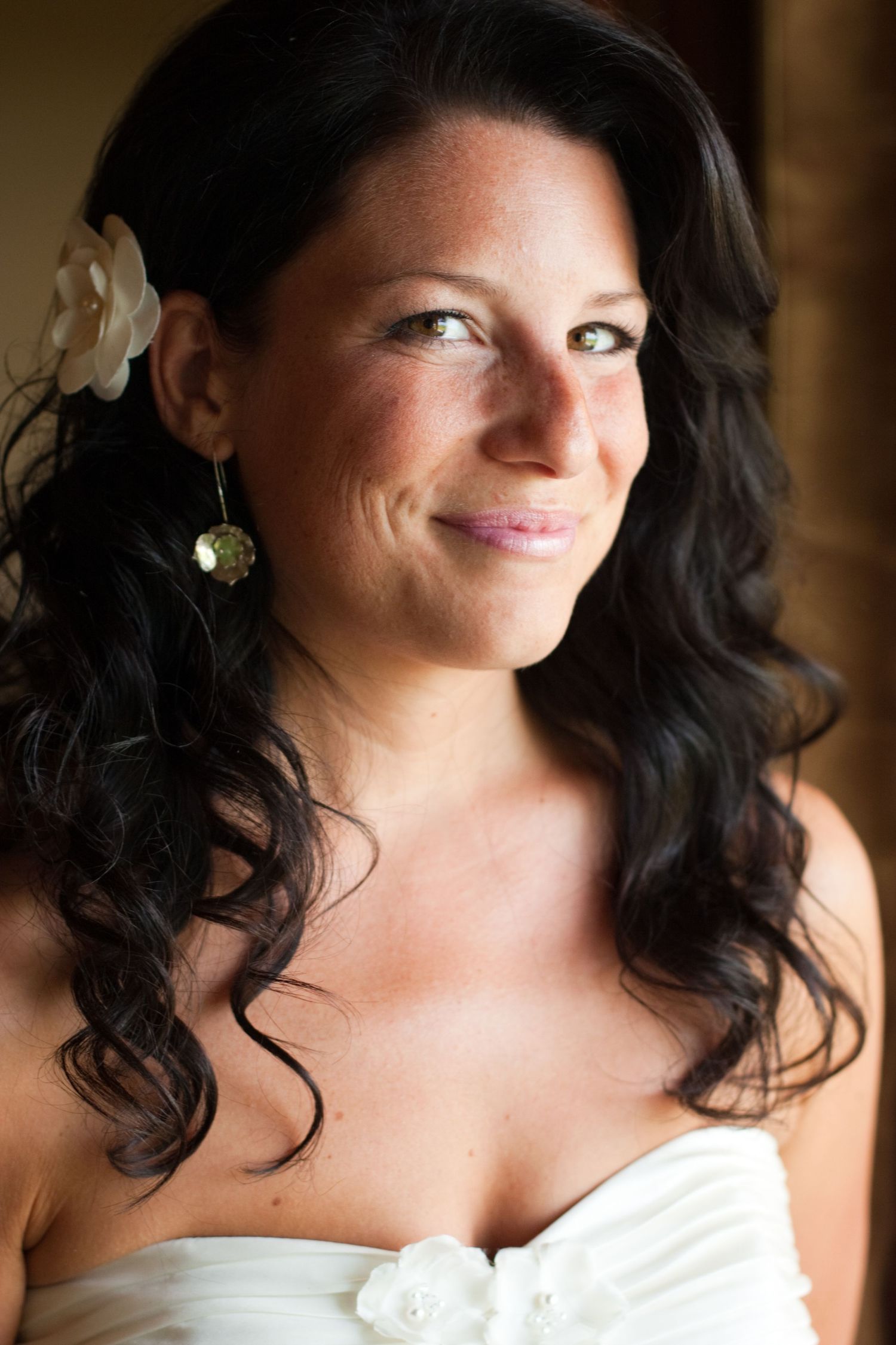 Portrait of bride with dark curly hair and white flower wearing strapless wedding dress.