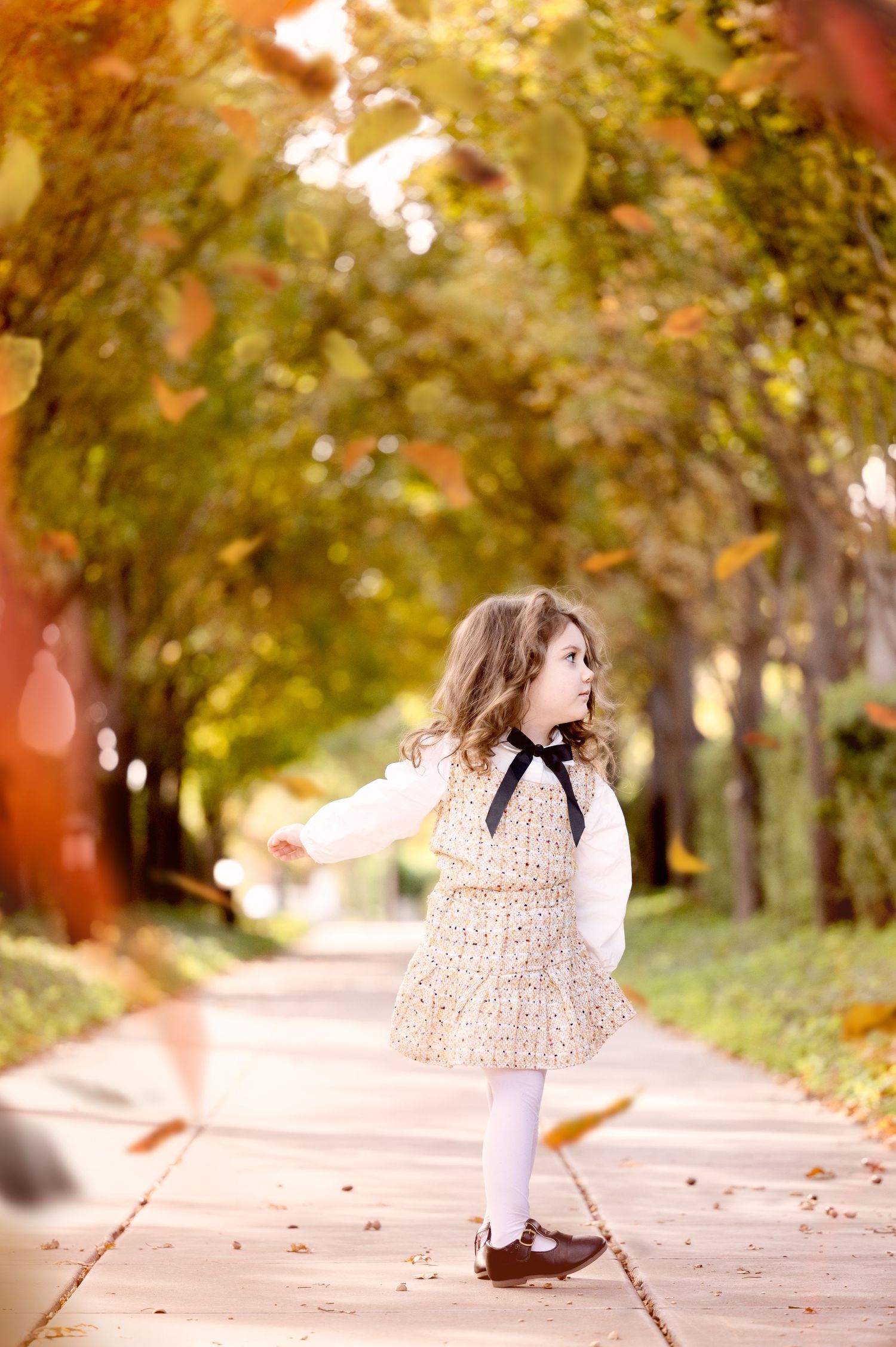 Child in peachy dress dances along a tree-lined path covered in autumn leaves.