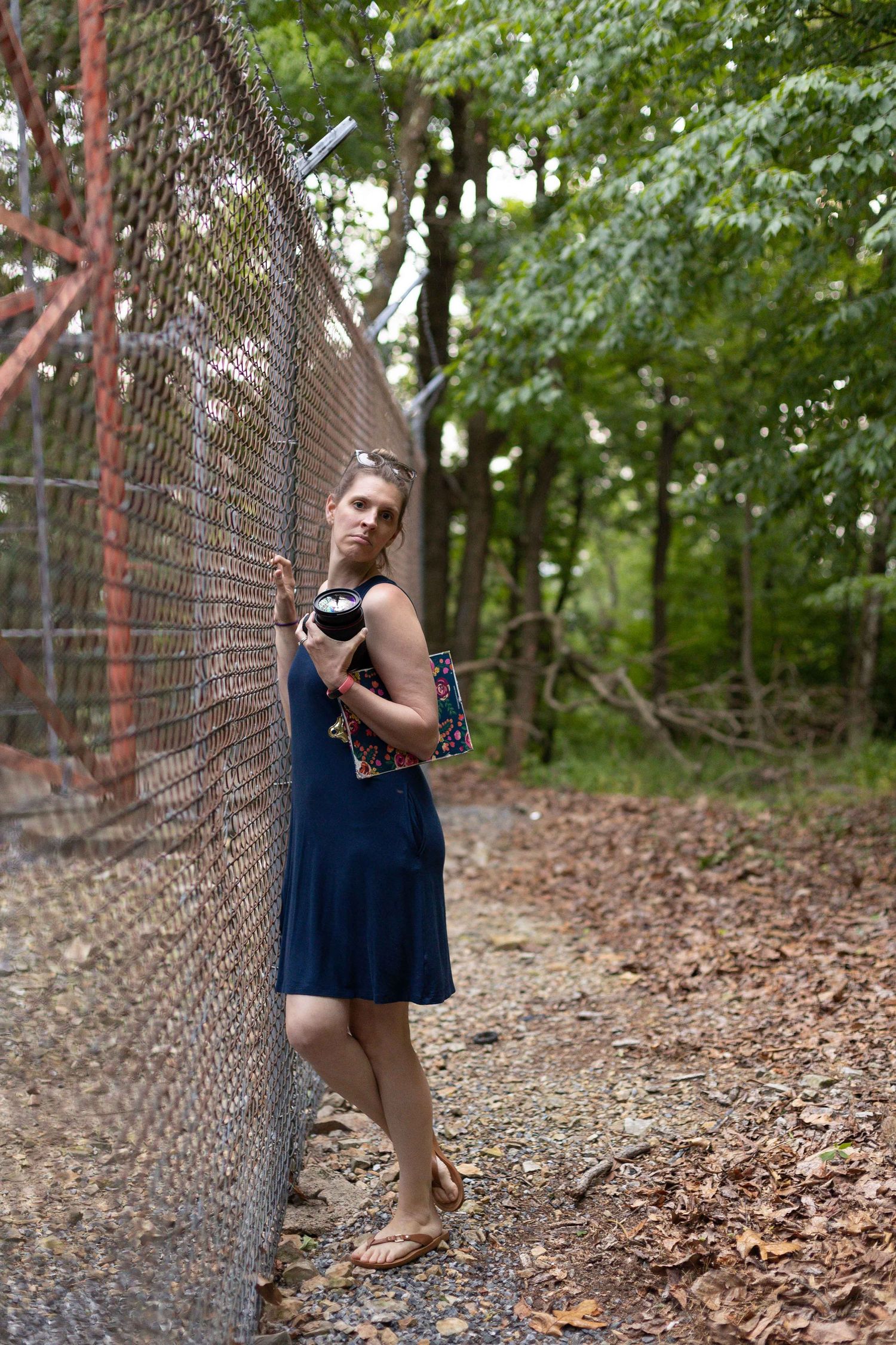 Someone in a navy dress poses against a chain-link fence in a wooded area.