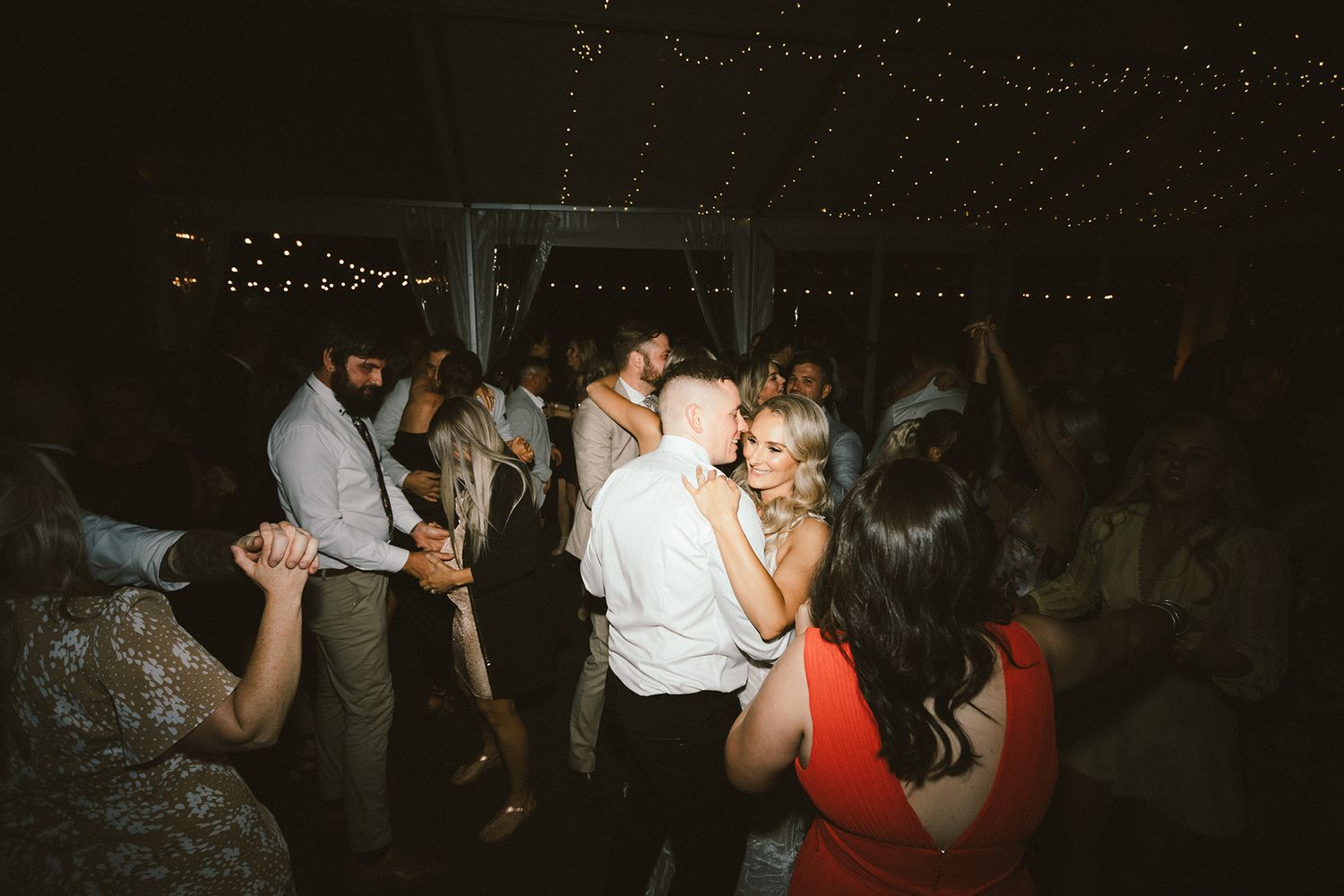 Wedding guests enjoying themselves on a dark dance floor illuminated by overhead string lights.
