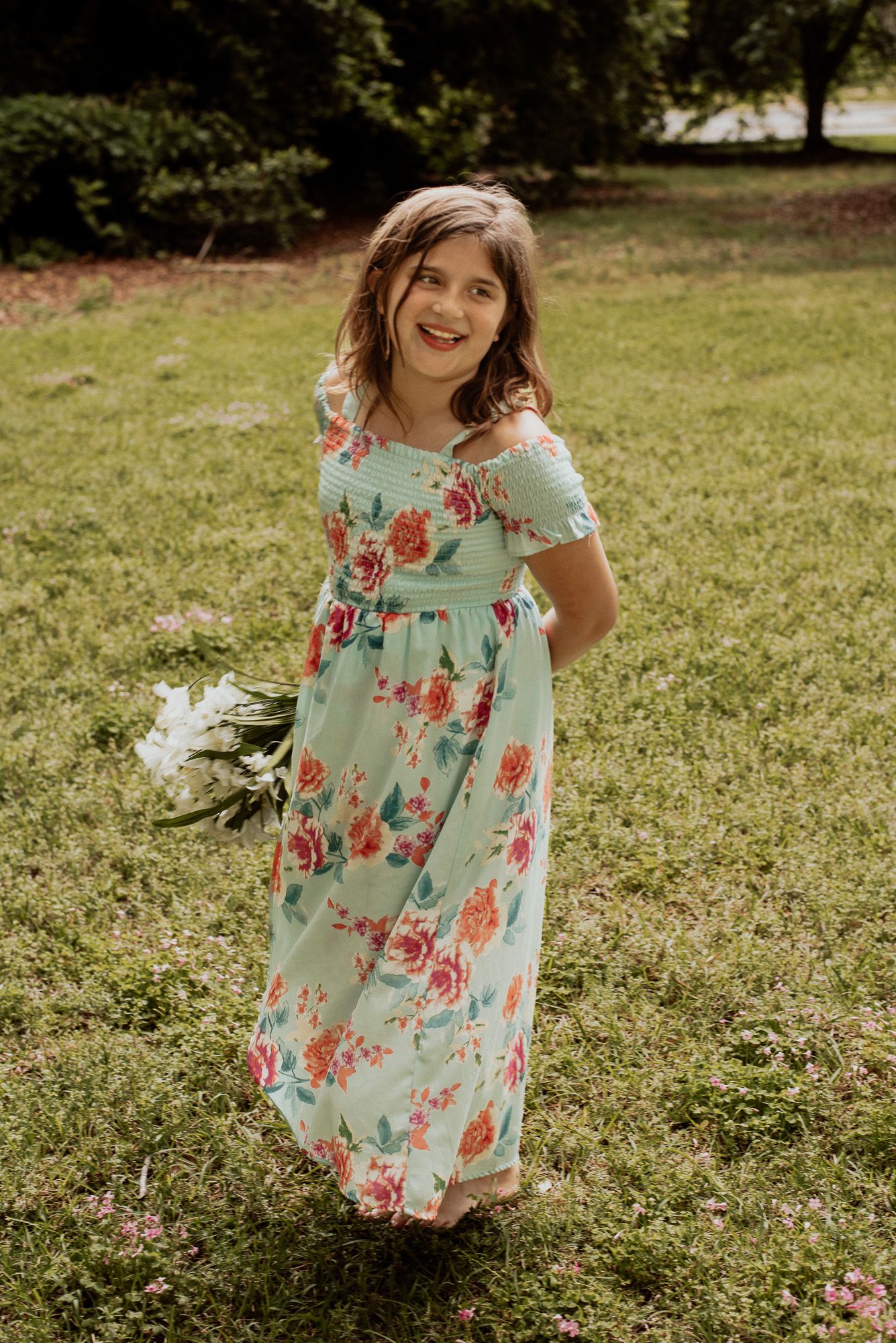 A person in a floral mint dress holds flowers while standing in a sunny garden.