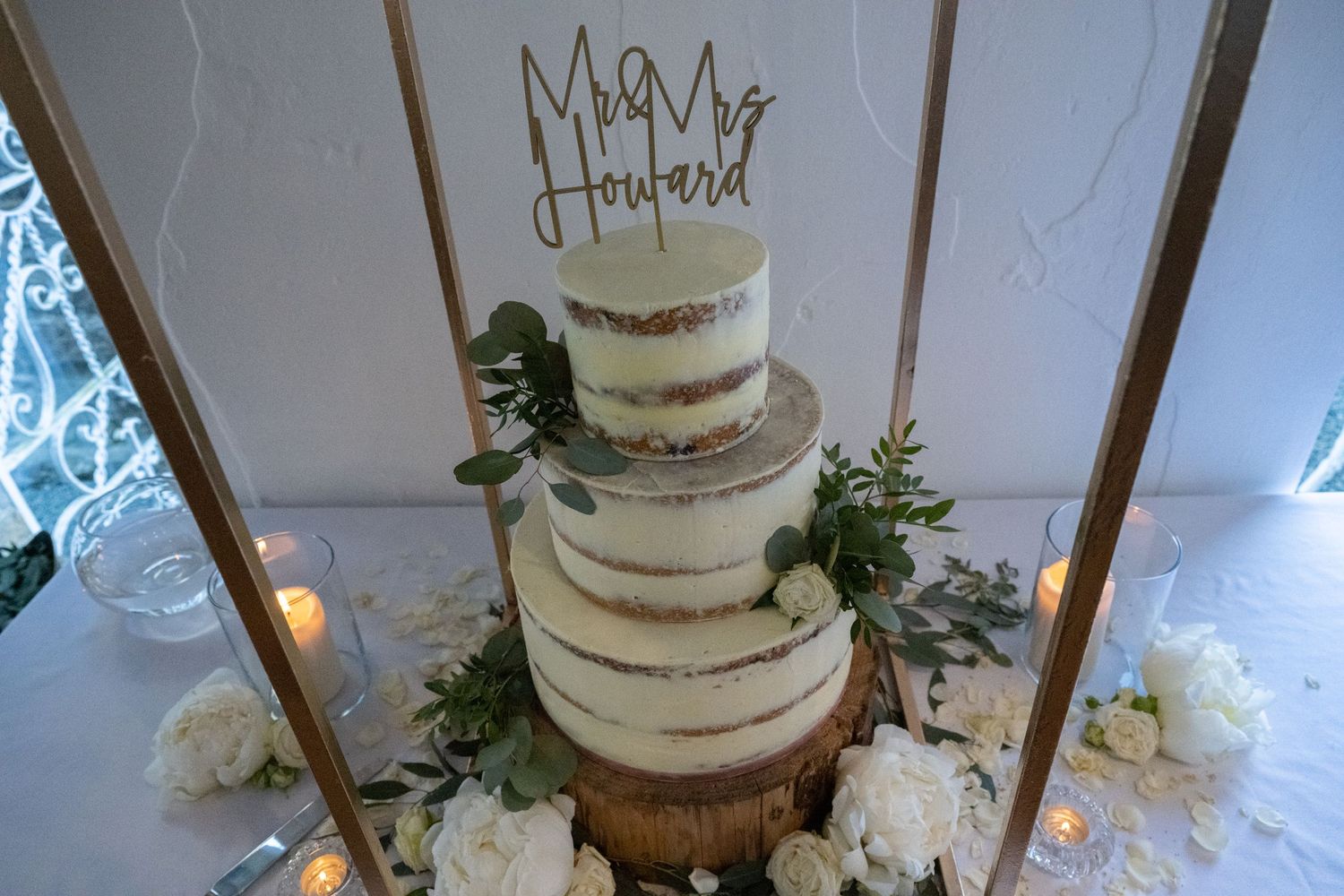 Three-tiered white semi-naked wedding cake with greenery decorations and a rustic topper.
