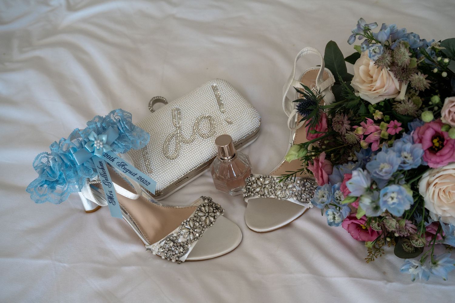 Wedding accessories including sparkly shoes, floral garter and colorful bouquet on white fabric.