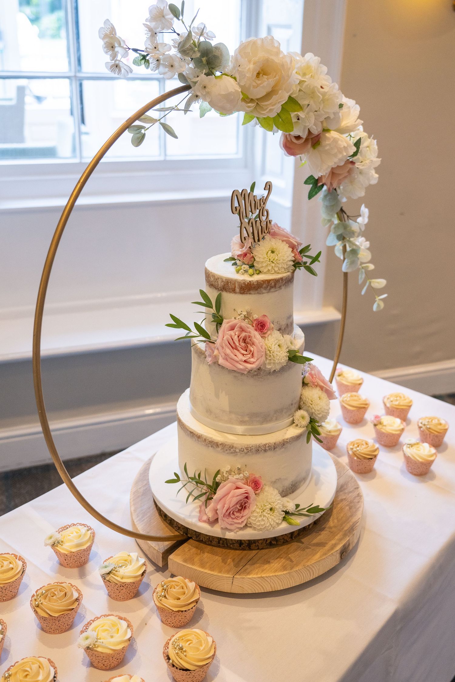Elegant three-tiered wedding cake with pink roses displayed on gold hoop stand with matching cupcakes.