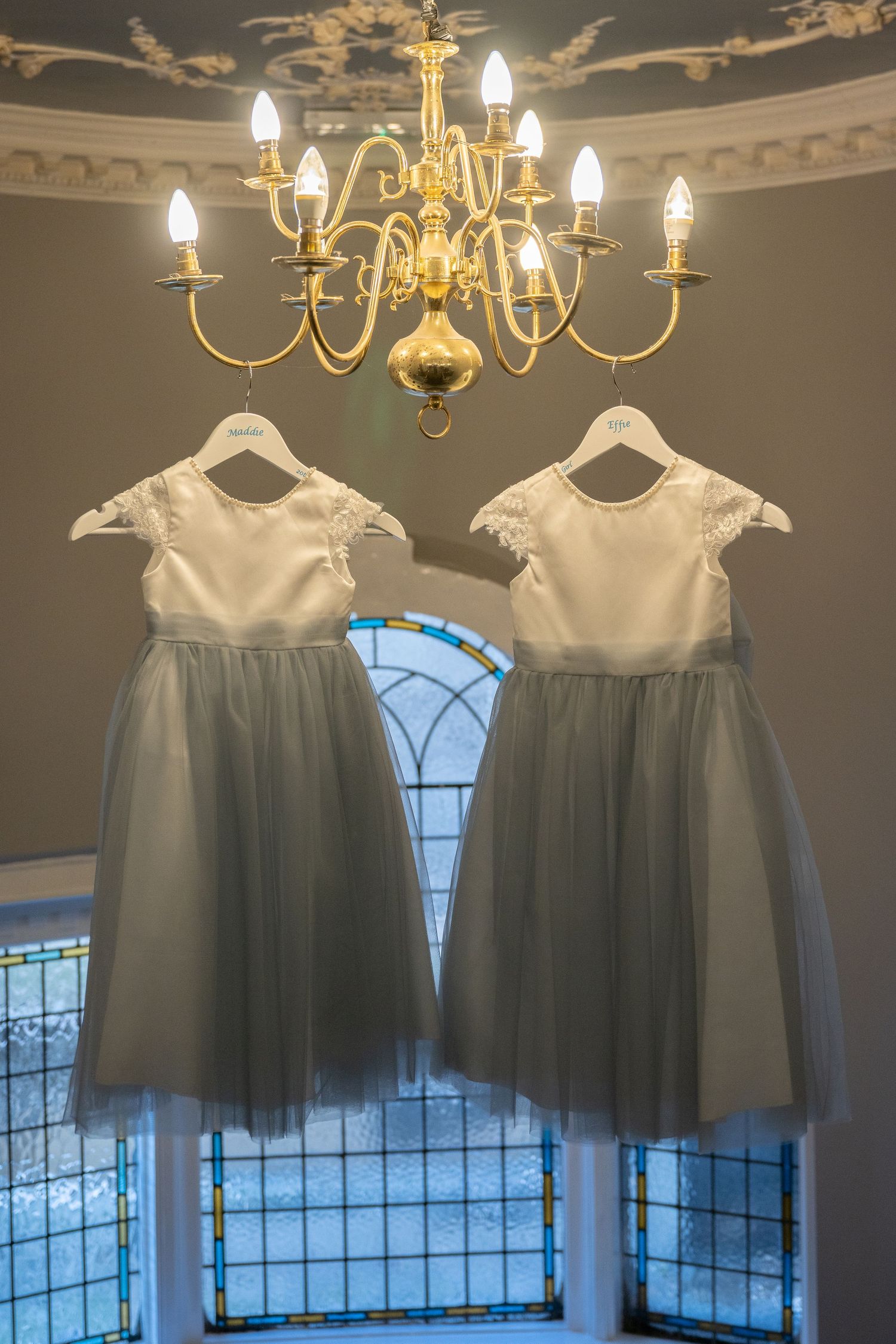 Two gray tulle flower girl dresses hanging beneath an elegant brass chandelier.