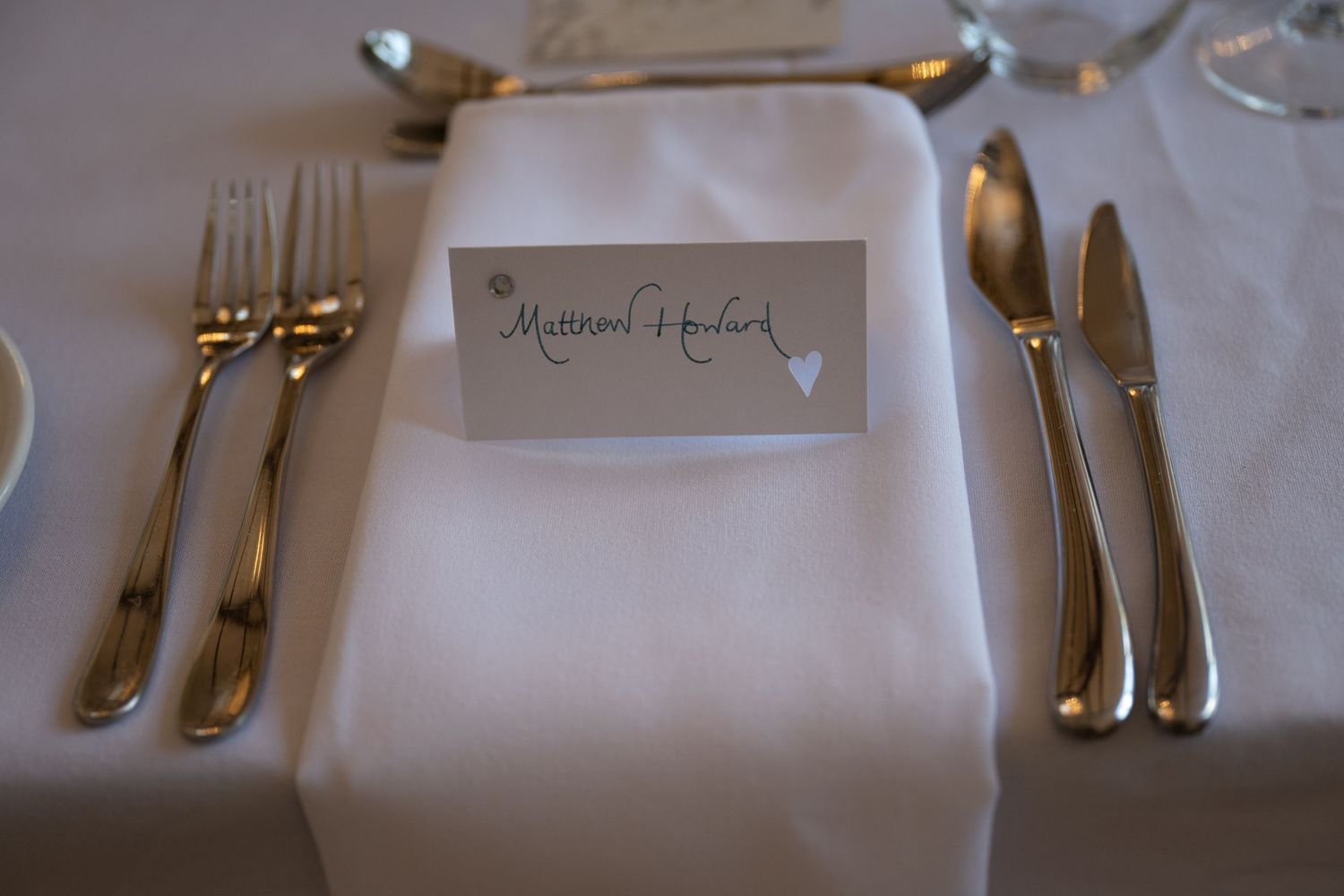 Elegant place setting with white napkin and name card surrounded by silver cutlery on formal dining table.