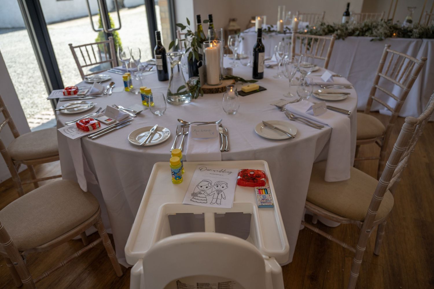 Wedding reception table settings with white linens and chiavari chairs in bright indoor venue.