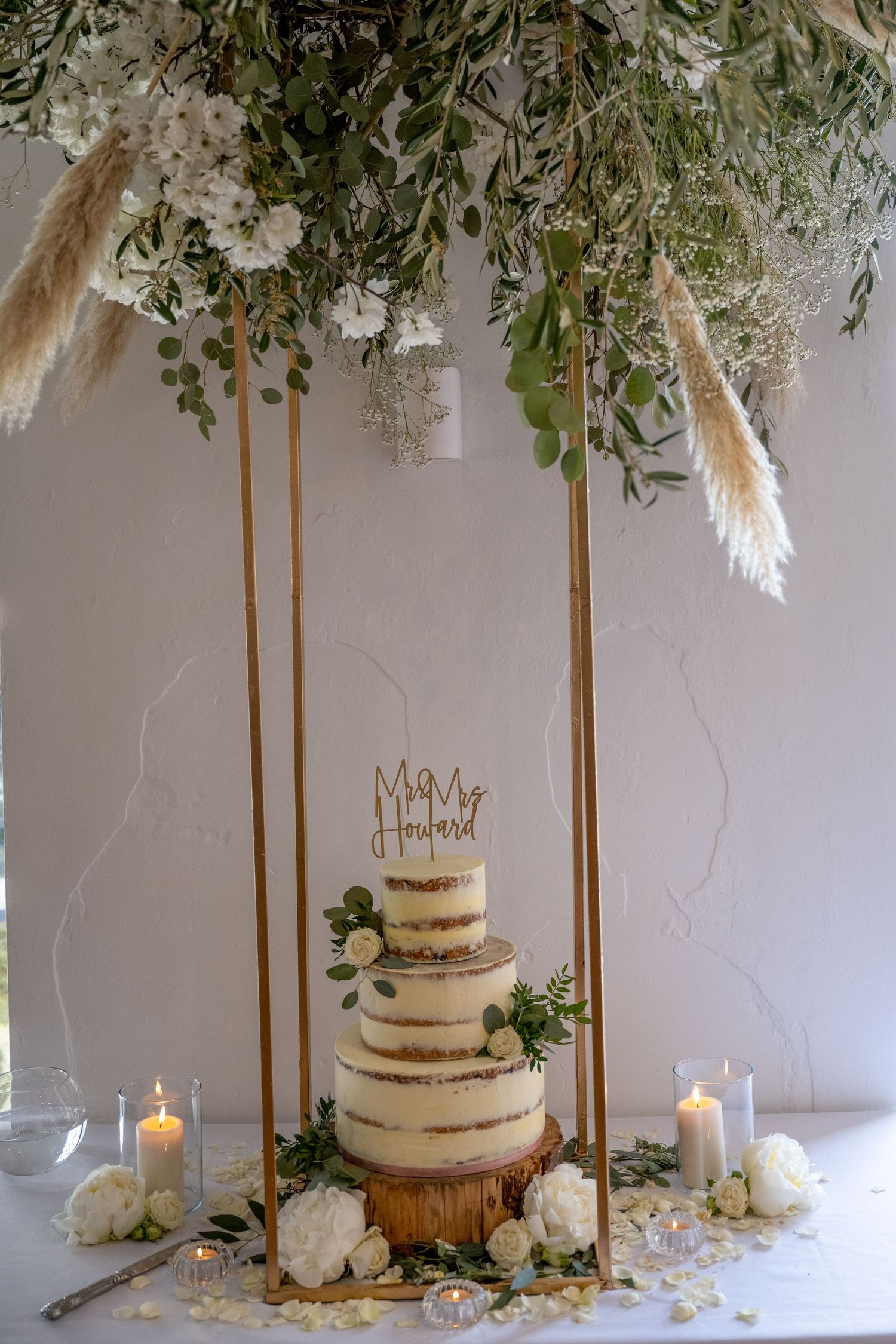 Rustic naked wedding cake displayed under hanging floral arrangement with candles and greenery.