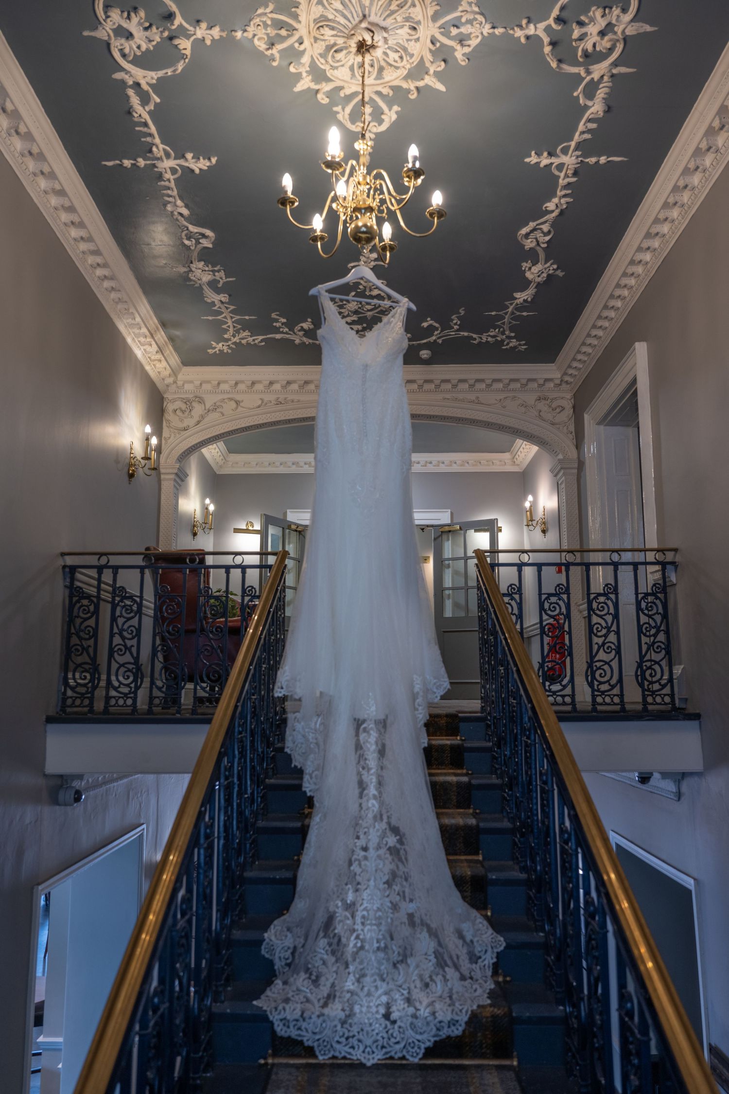 Lace wedding dress hanging in grand stairwell with ornate ceiling and chandelier.