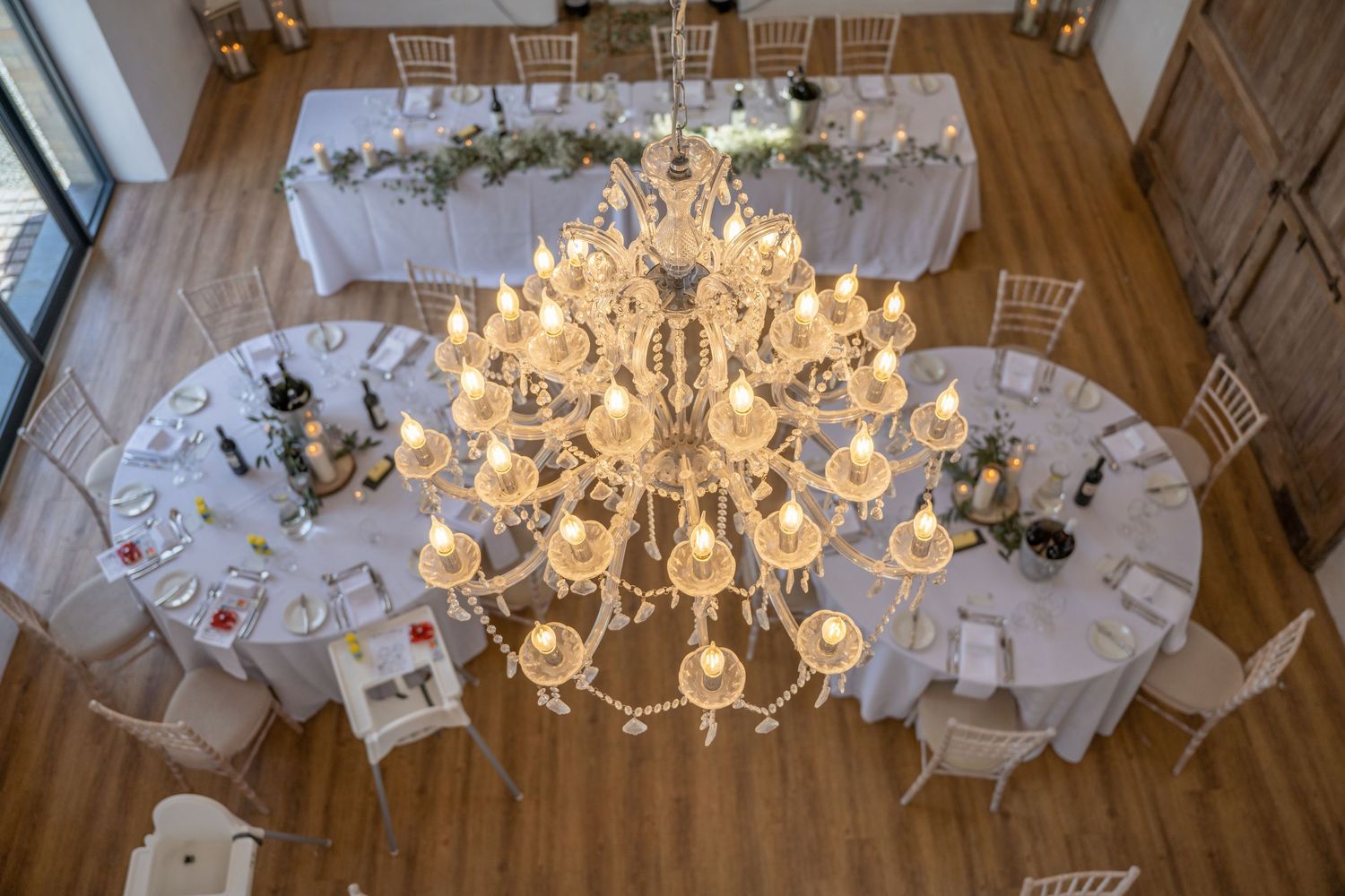 Elegant wedding reception setup with round tables and a grand crystal chandelier in a wooden-floored venue.