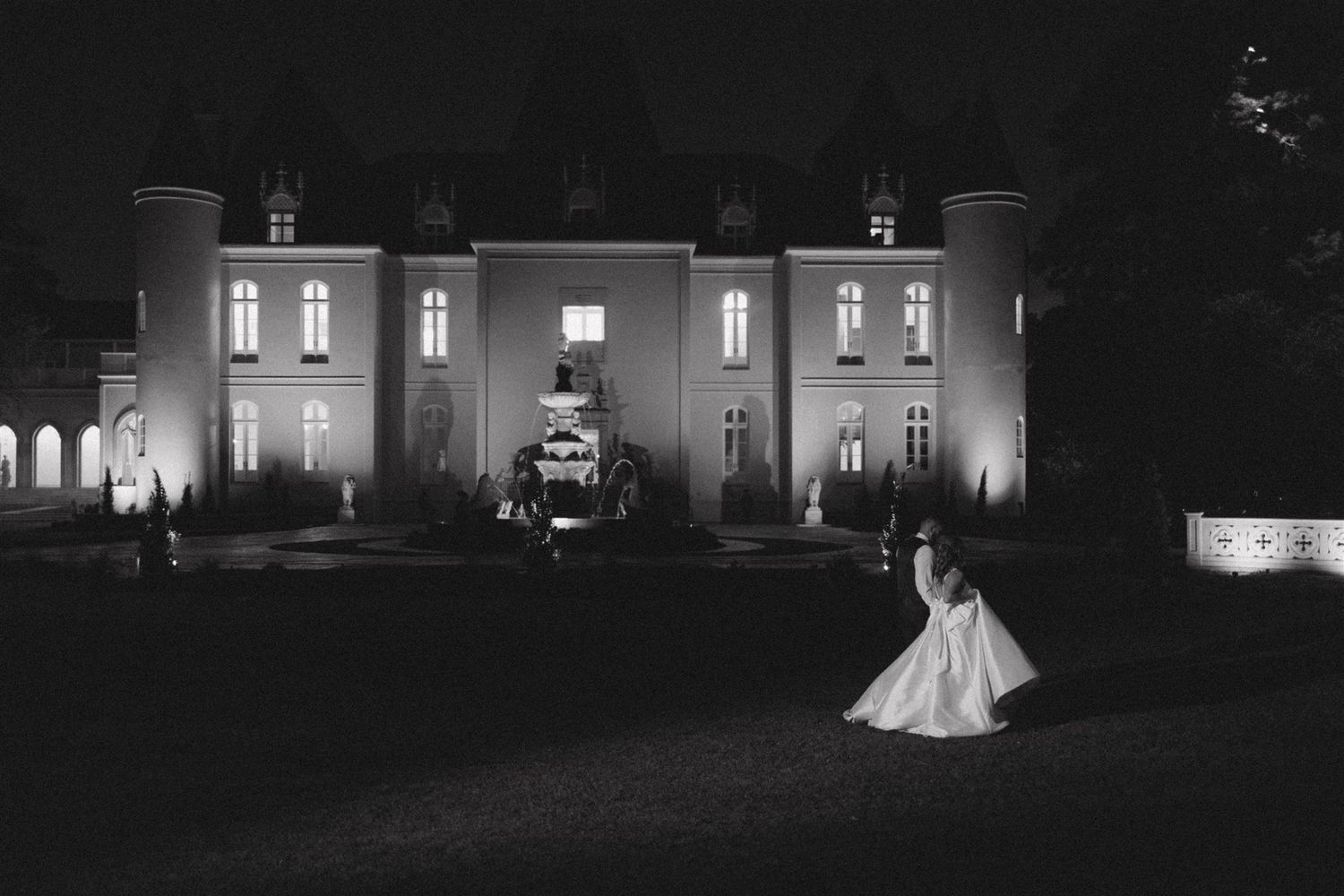 A romantic black and white night scene shows a couple dancing on a lawn in front of an illuminated manor house.