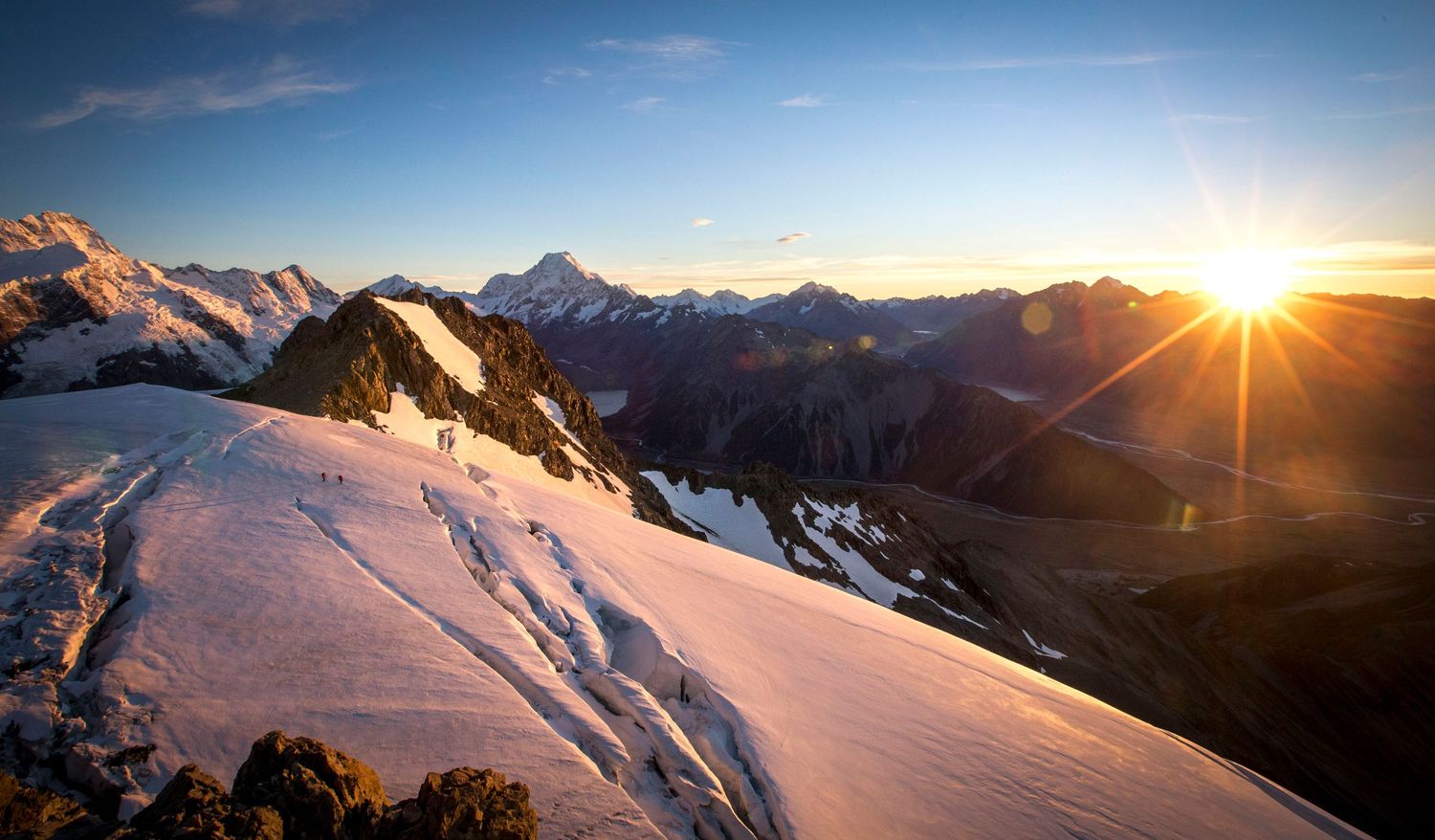 Breathtaking sunrise over snow-covered mountain peaks with a sunburst and vast alpine valley below.