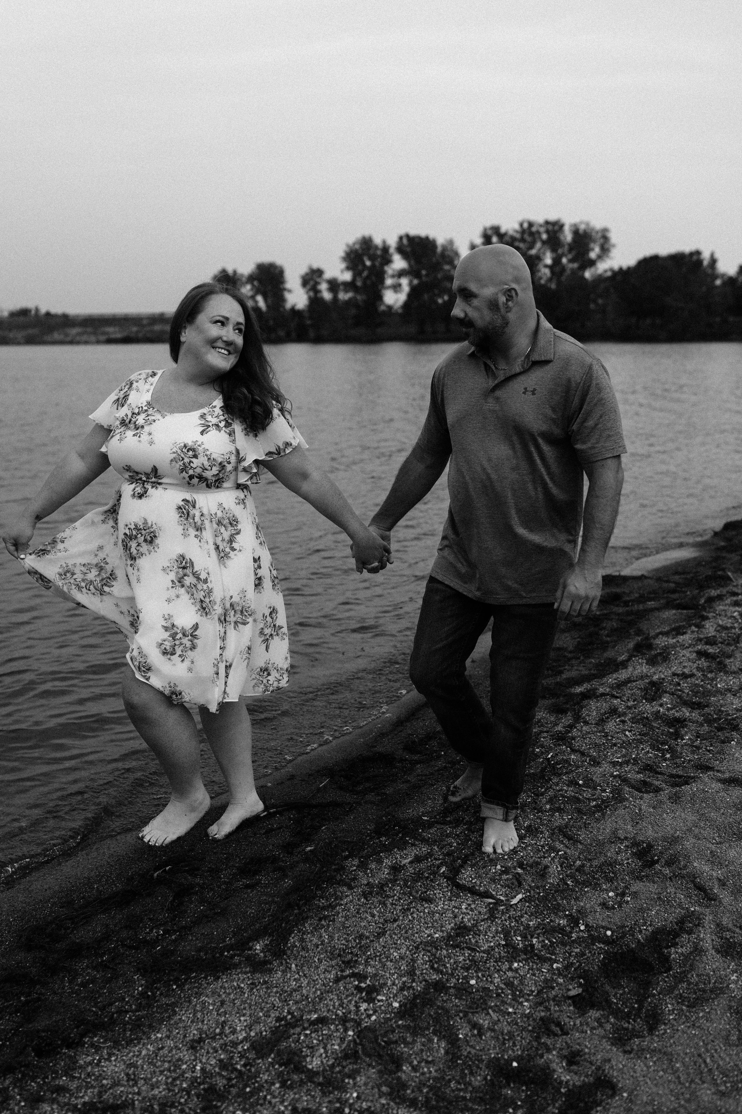 Couple holding hands while walking barefoot along lakeside in black and white.