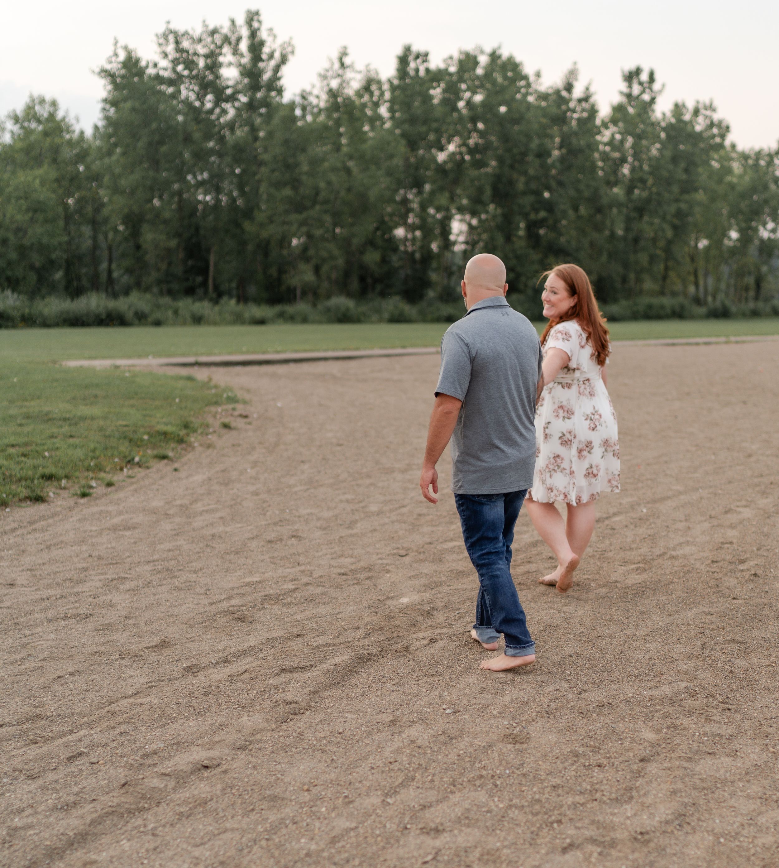 Two people walk hand in hand on a dirt path surrounded by trees at sunset.
