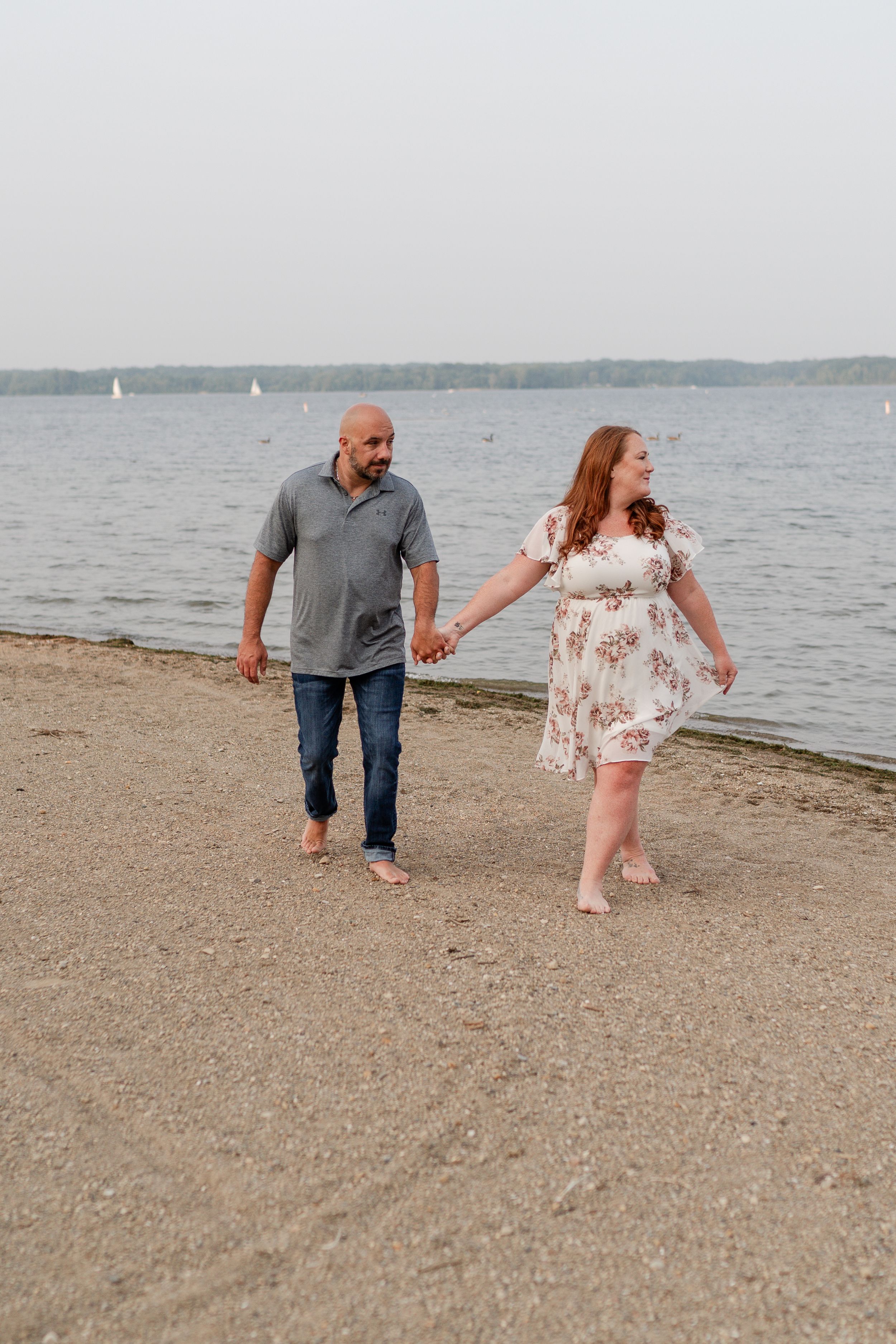 A couple holding hands while strolling along a beach with sailboats in the distance.