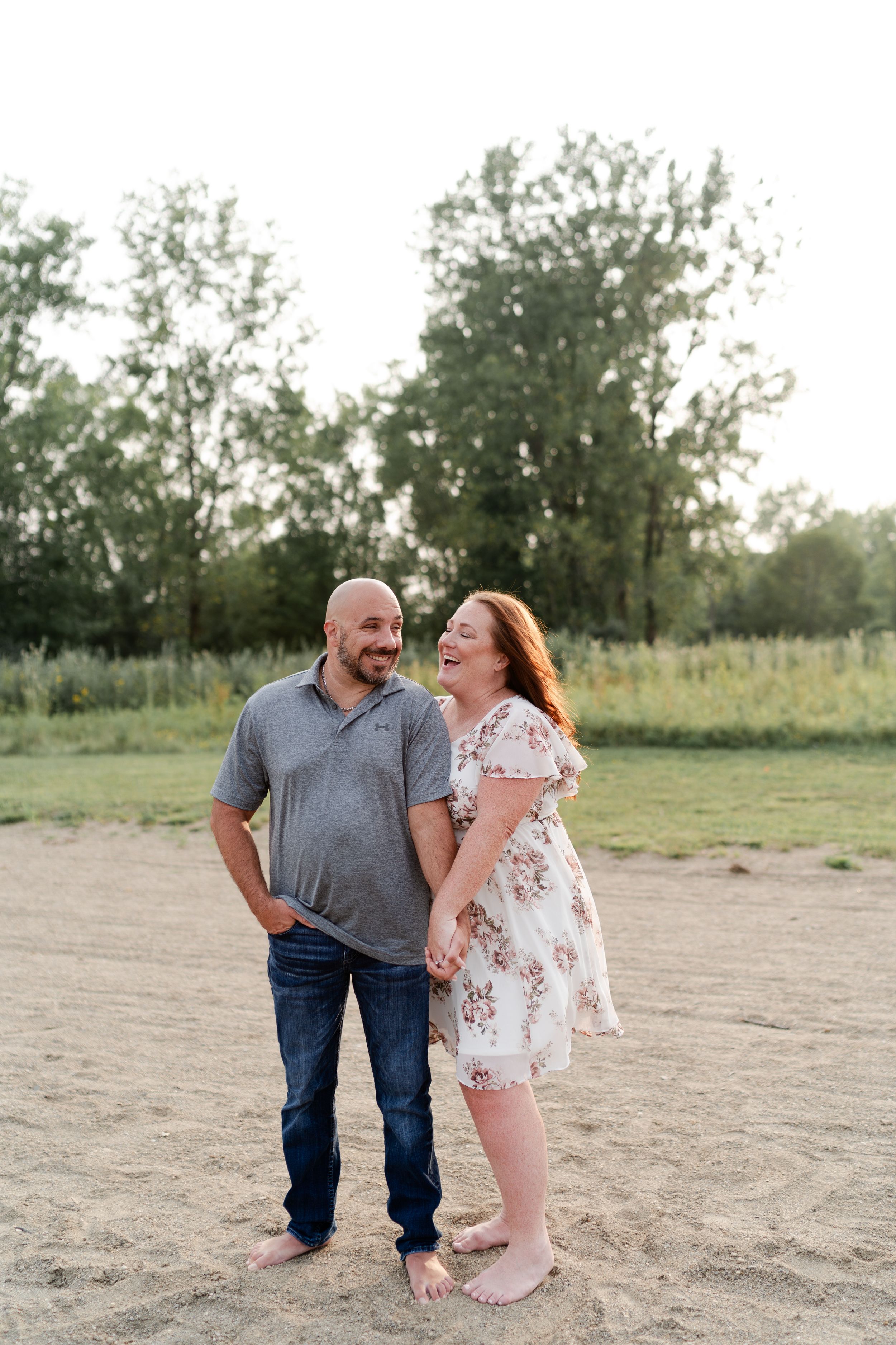 A couple standing barefoot together on a sandy beach with trees in the background.