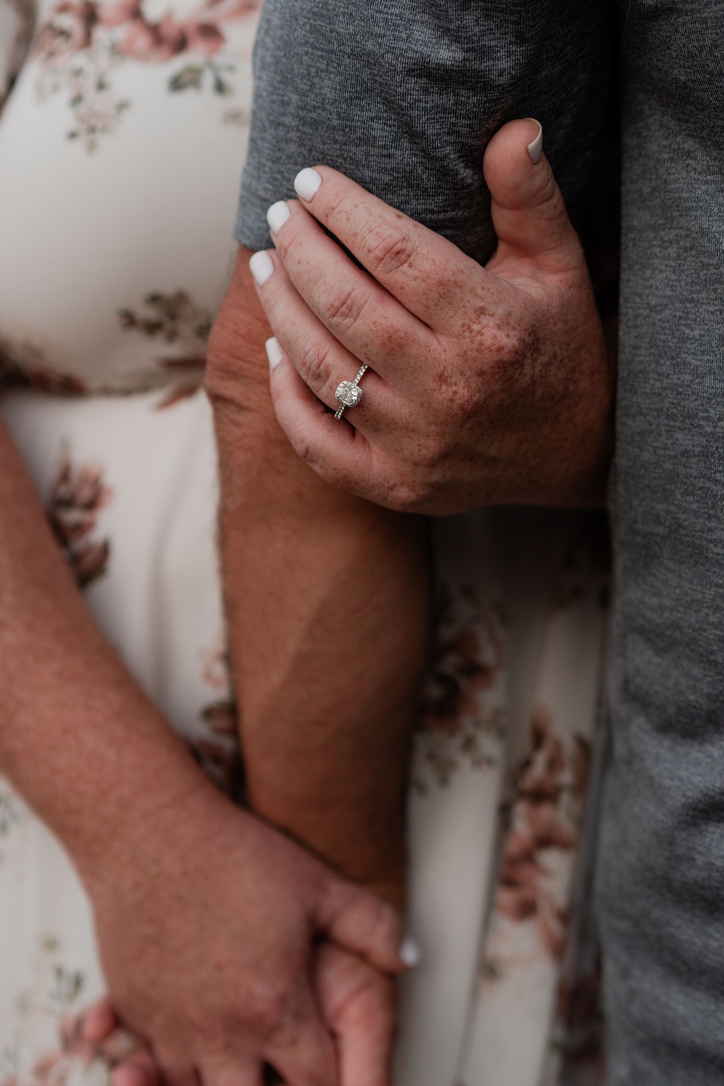 Close-up of hands with engagement ring against floral dress and gray shirt background.