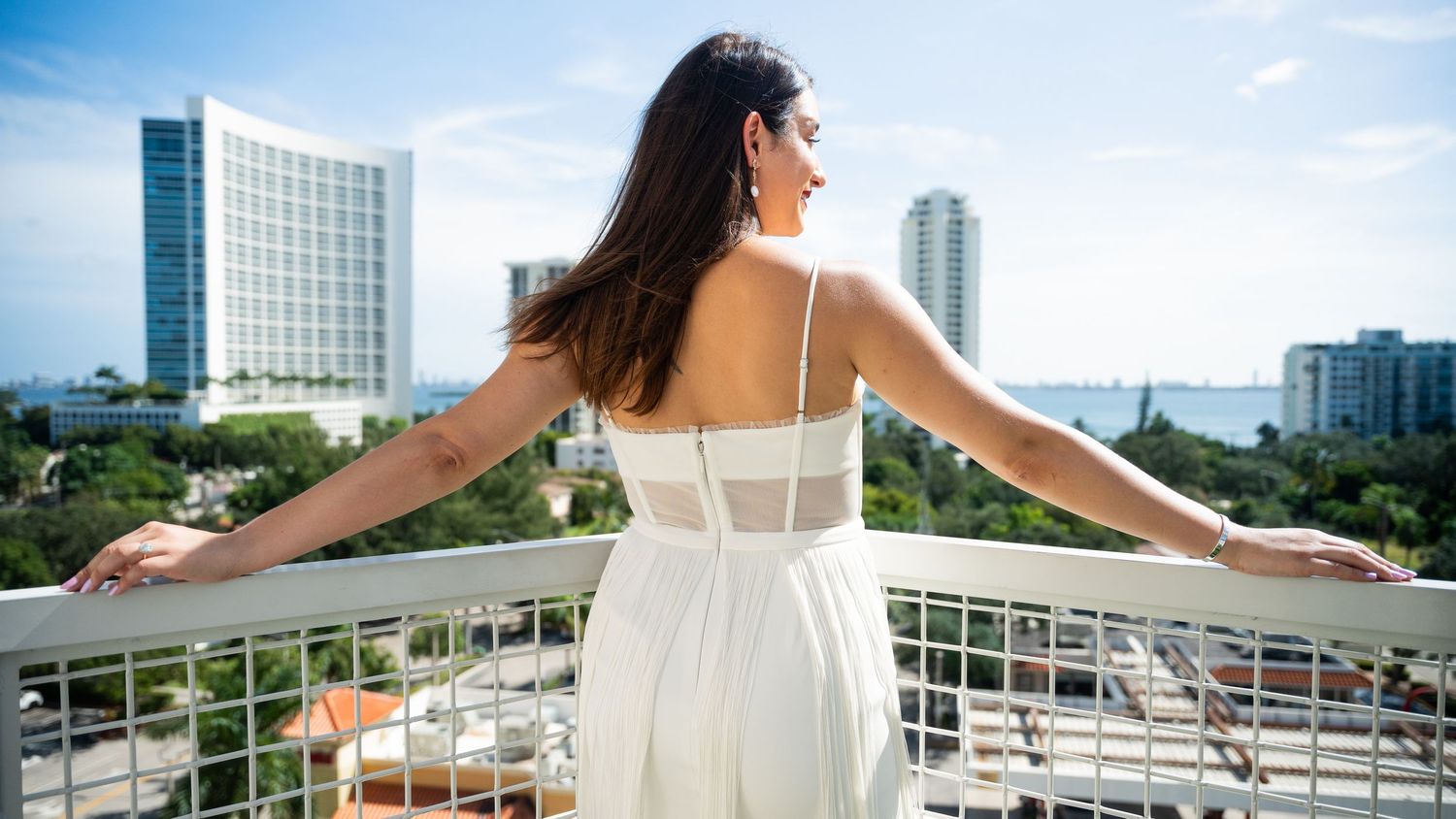 Woman in white dress stands with arms outstretched on balcony overlooking city skyline.