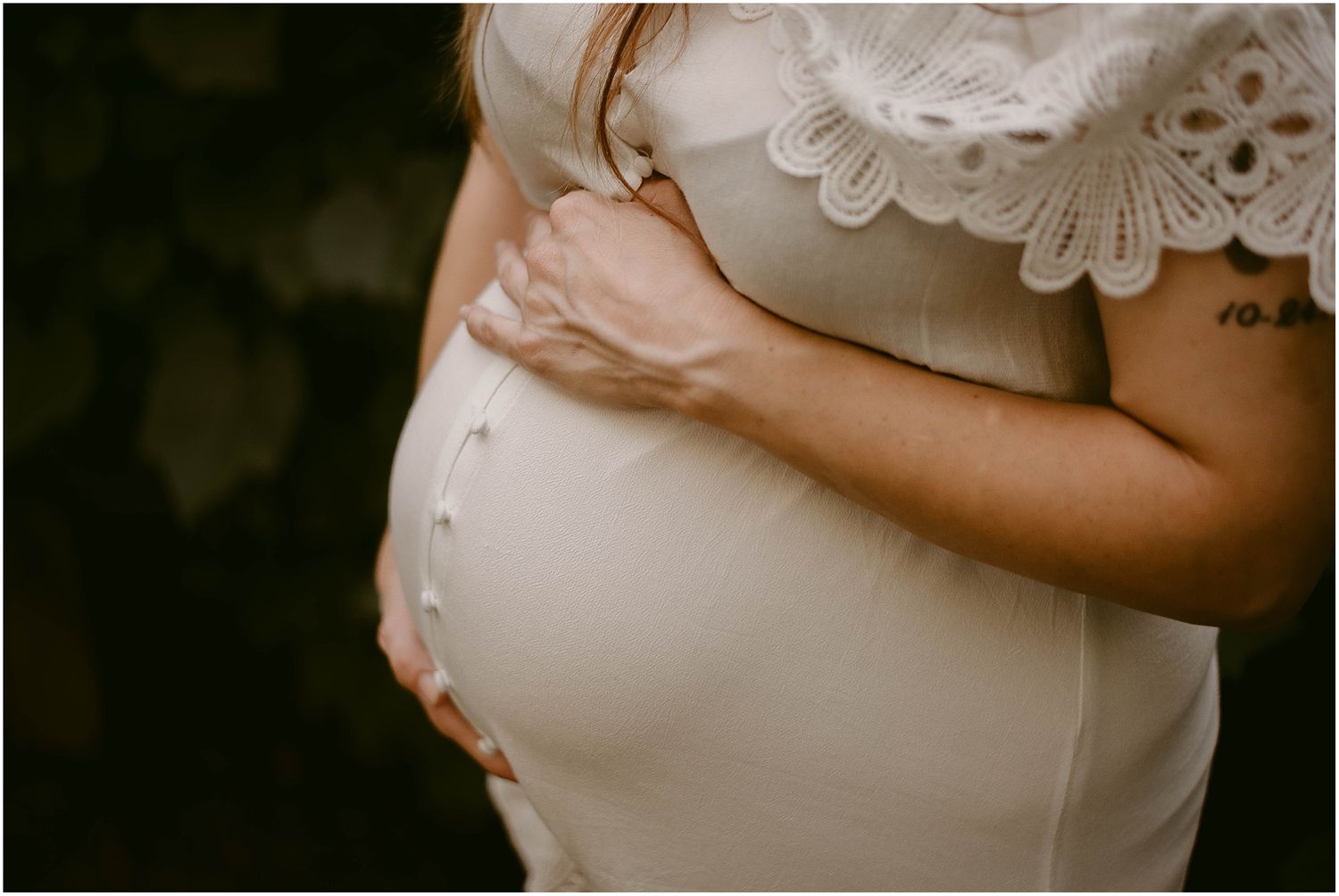 Close-up detail shot of pregnant belly in white lace dress with hands gently cradling.